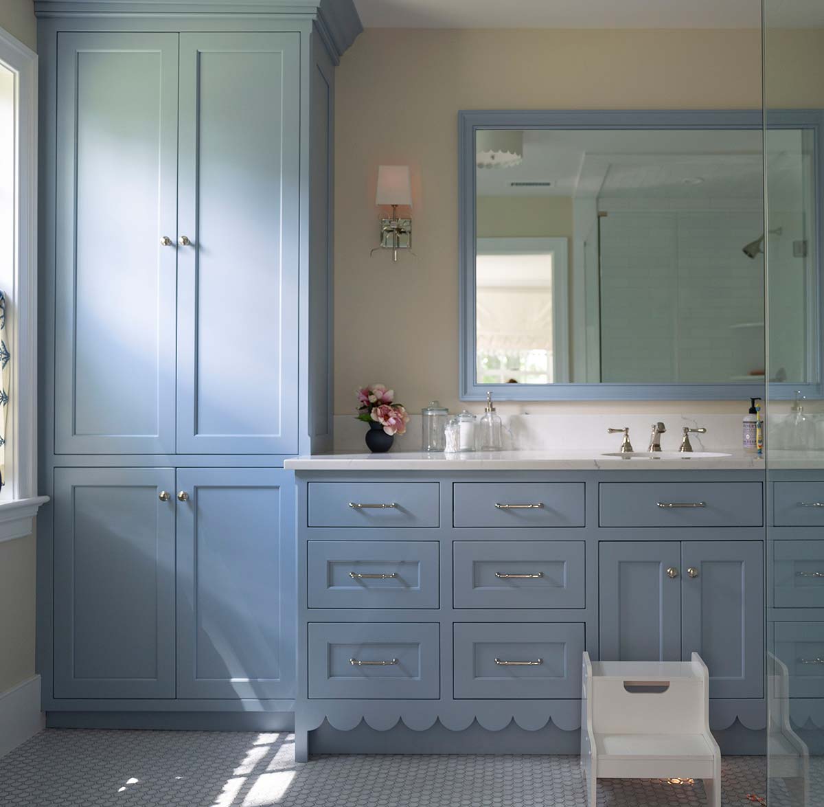 Blue bathroom cabinetry with scalloped edges, large mirror, and sink. Features a vase of flowers, white stool, and tiled floor with natural light.