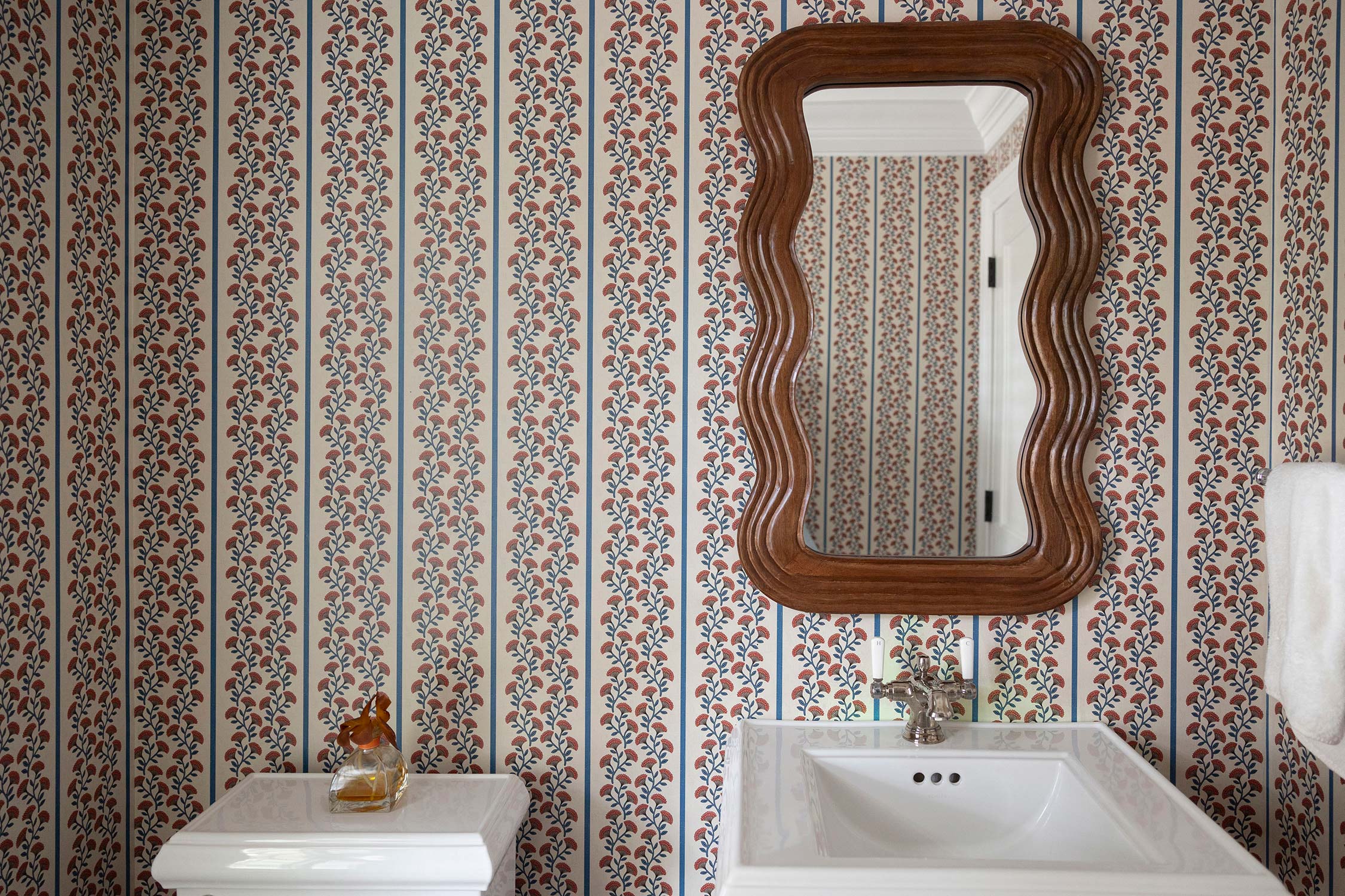 A chic bathroom with floral-patterned wallpaper, a wooden wavy mirror, white sink, and a small table with a bottle.