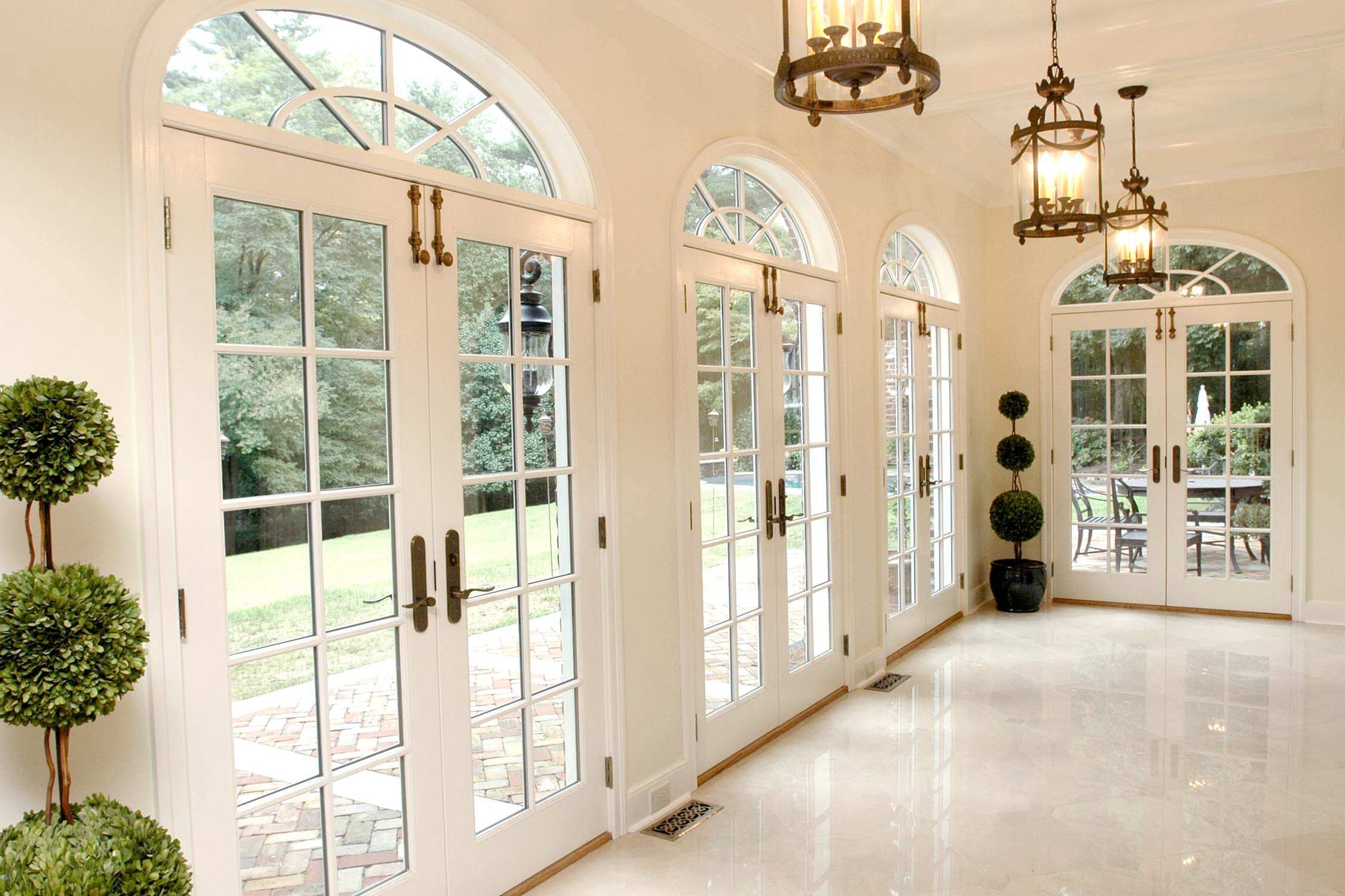 Elegant sunroom hallway with arched white French doors, hanging lanterns, topiary plants, polished marble floors, and views of lush outdoor patio.