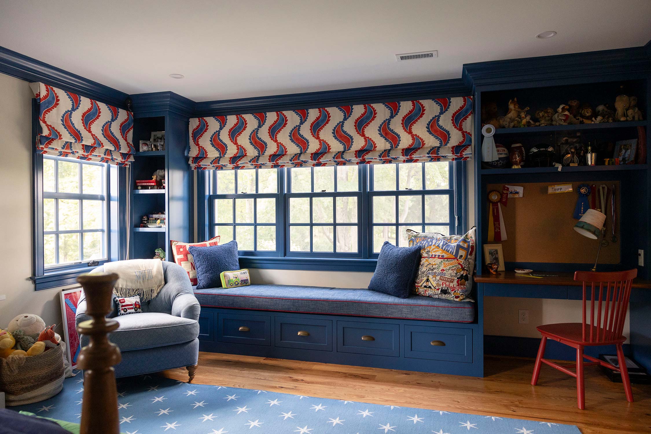 Child's room with blue window seat, colorful cushions, and red chair. Features patterned curtains, star rug, and shelves with toys on wood floor.