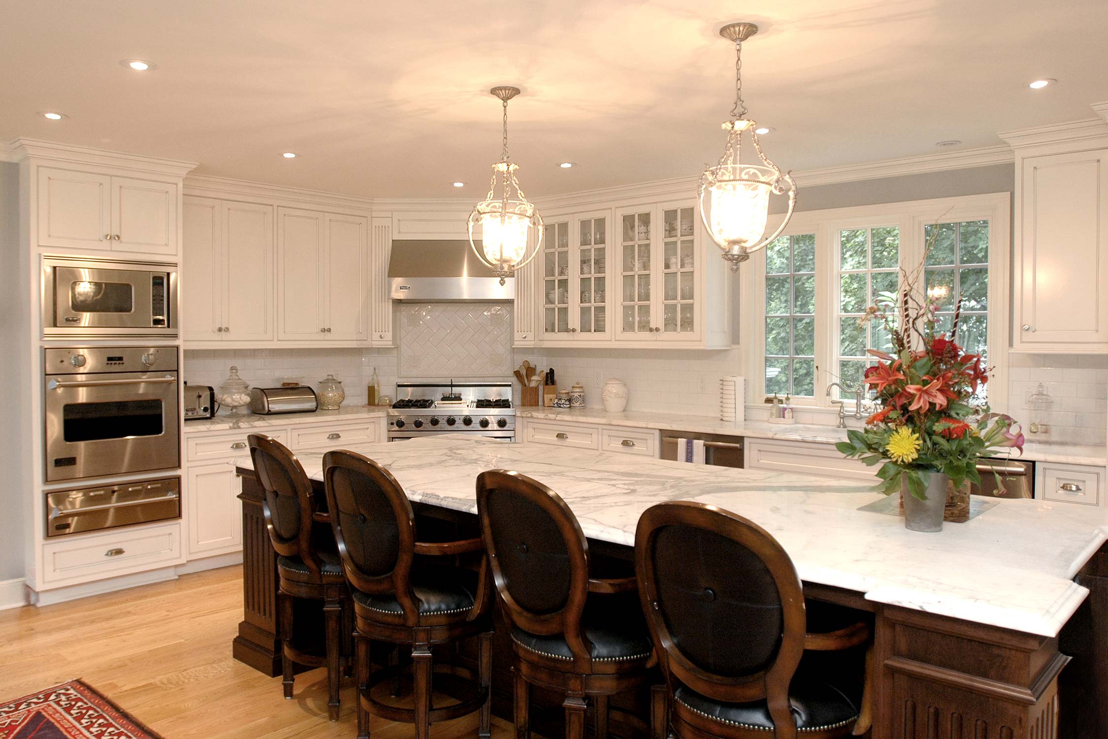 Luxurious kitchen with white cabinets, marble island bar, dark wood stools, stainless appliances, pendant lights, and vibrant floral centerpiece.