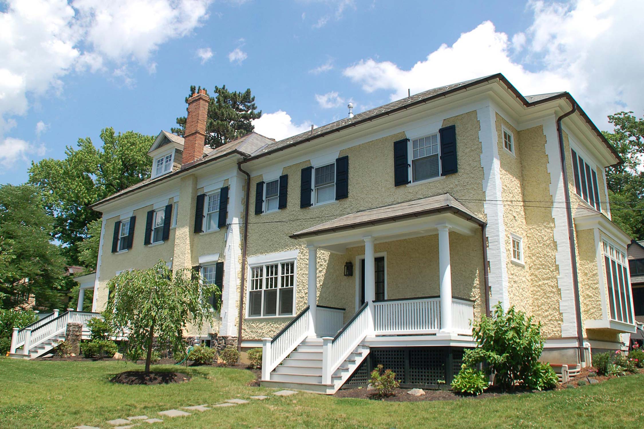 Charming yellow house with black shutters, white porch, and steps, surrounded by lush greenery under a bright blue sky.