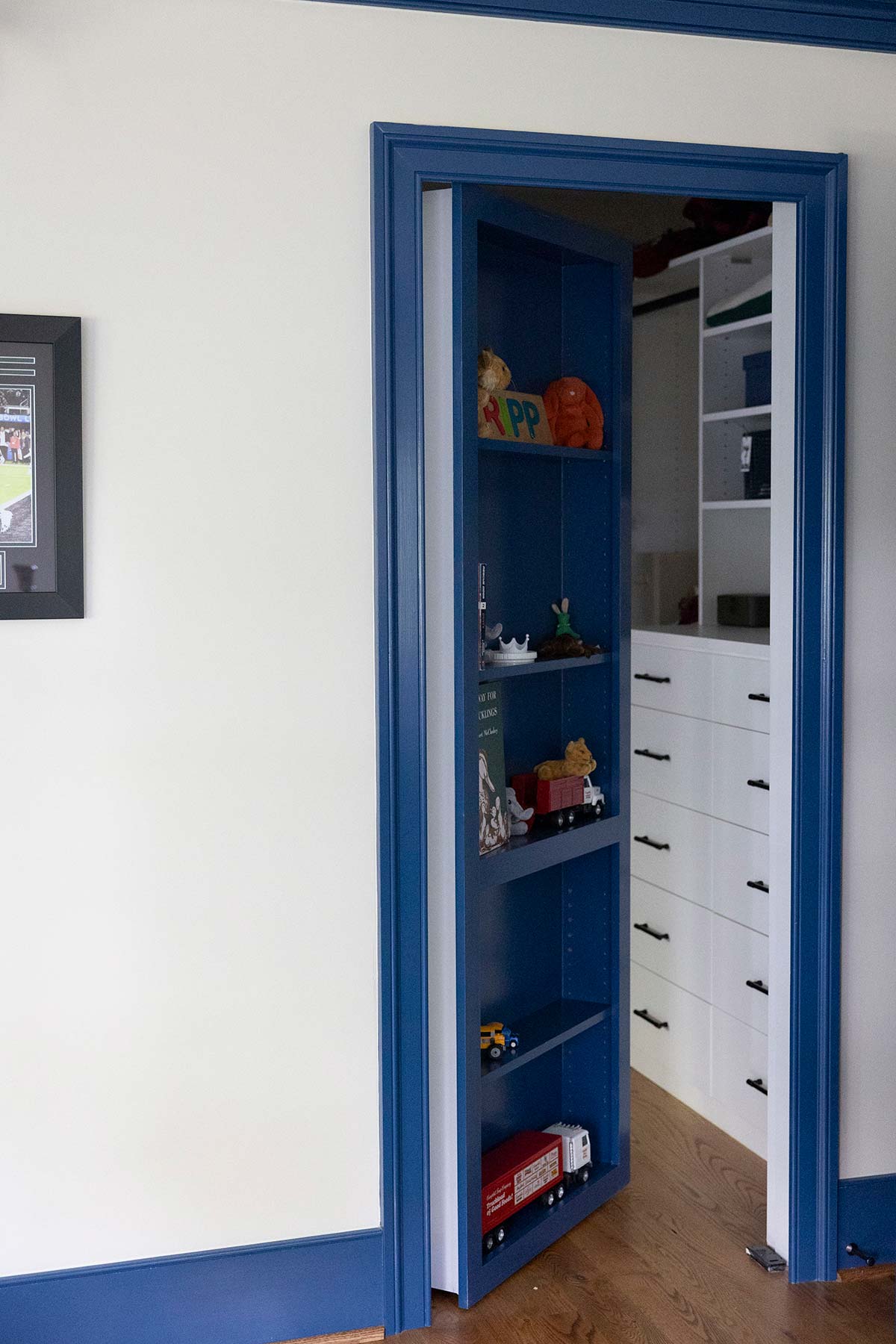 Blue bookshelf door revealing a closet with white drawers and toys. Includes a framed picture and wooden floor in a bright room.