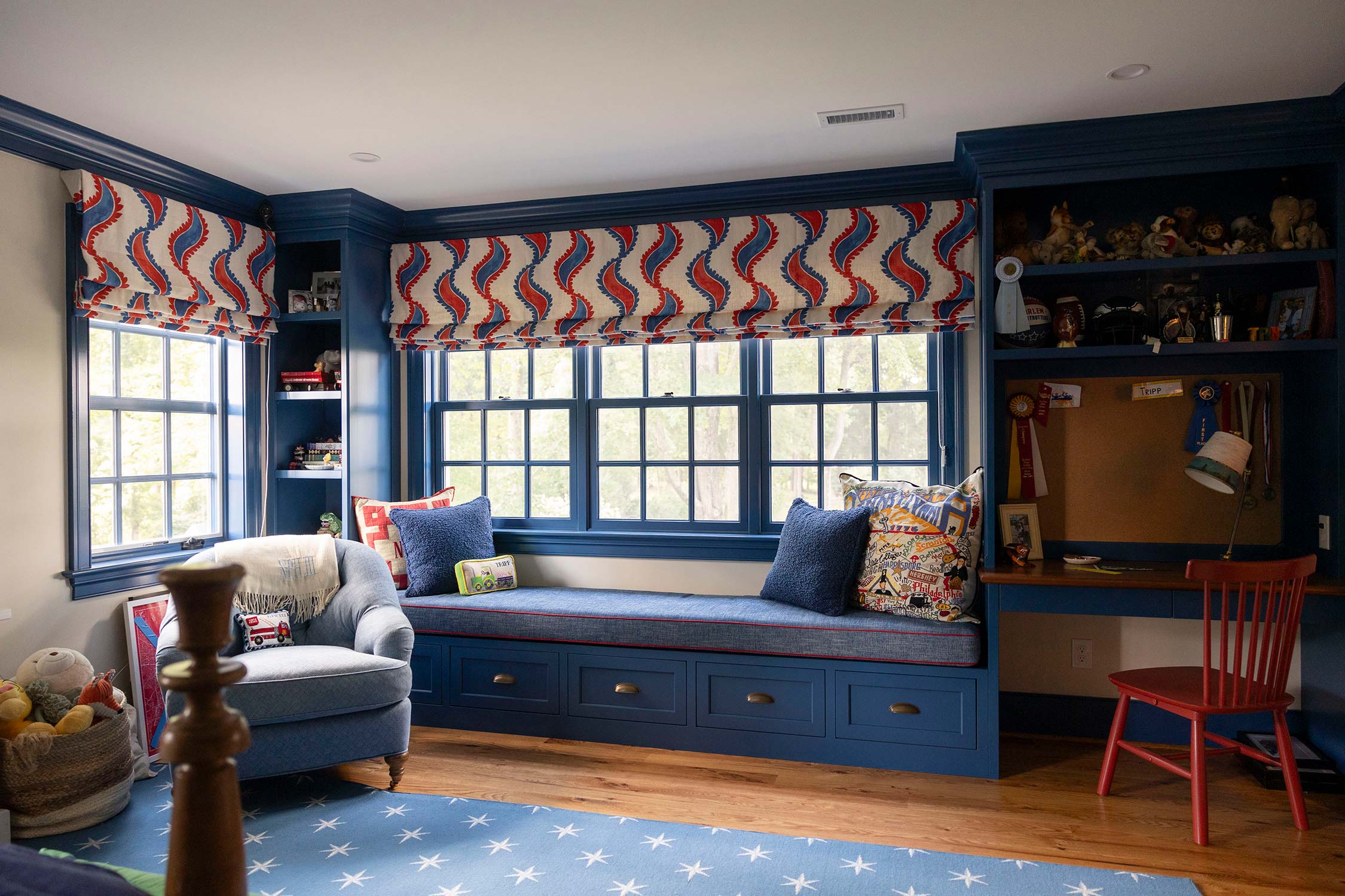 Child's room with blue window seat, colorful cushions, and red chair. Features patterned curtains, star rug, and shelves with toys on wood floor.