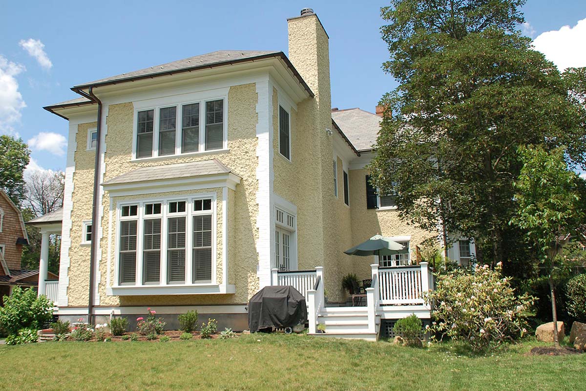 Cozy back view of yellow stucco house with bay windows, white deck, covered grill, umbrella, and blooming garden under blue skies.