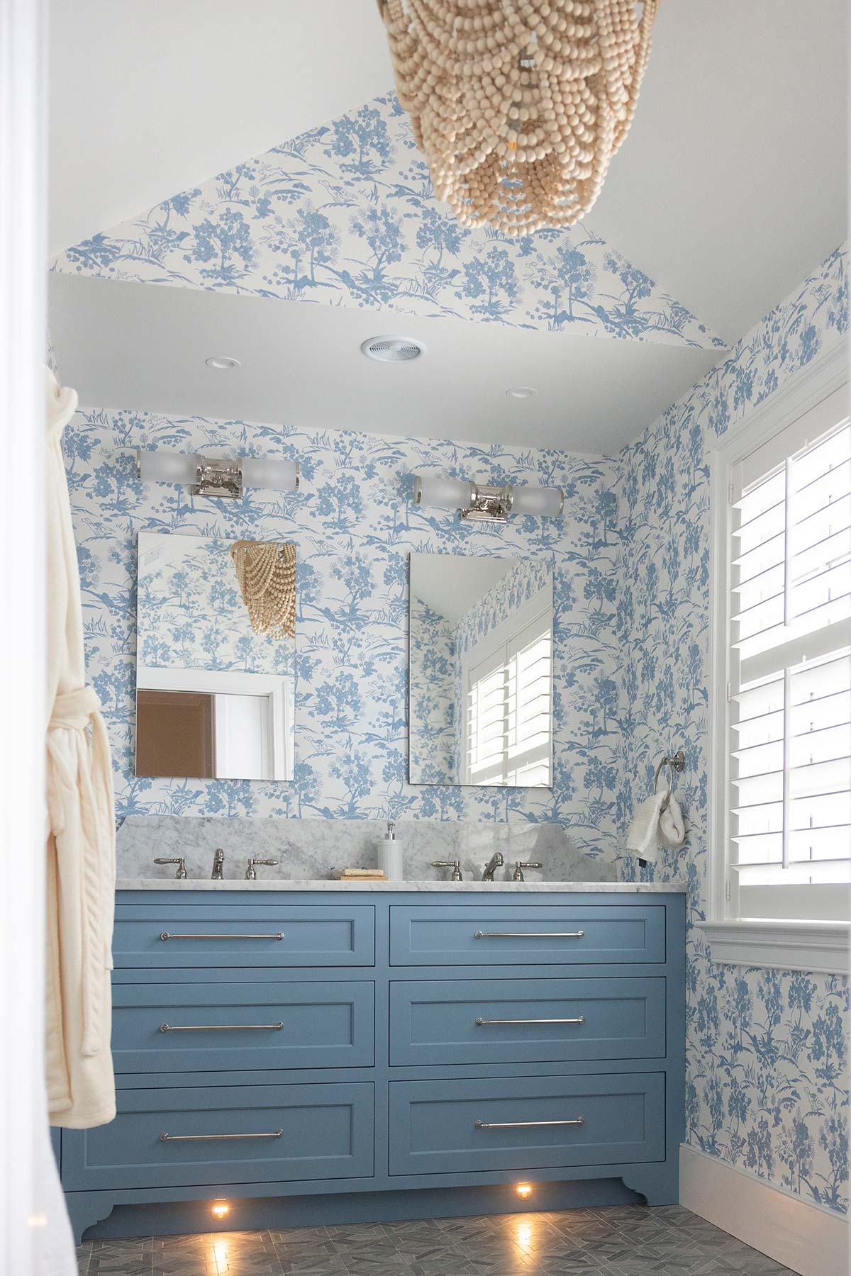 Blue bathroom with floral wallpaper, double sink, and beaded chandelier. Features mirrors, under-cabinet lighting, and tiled floor with natural light.