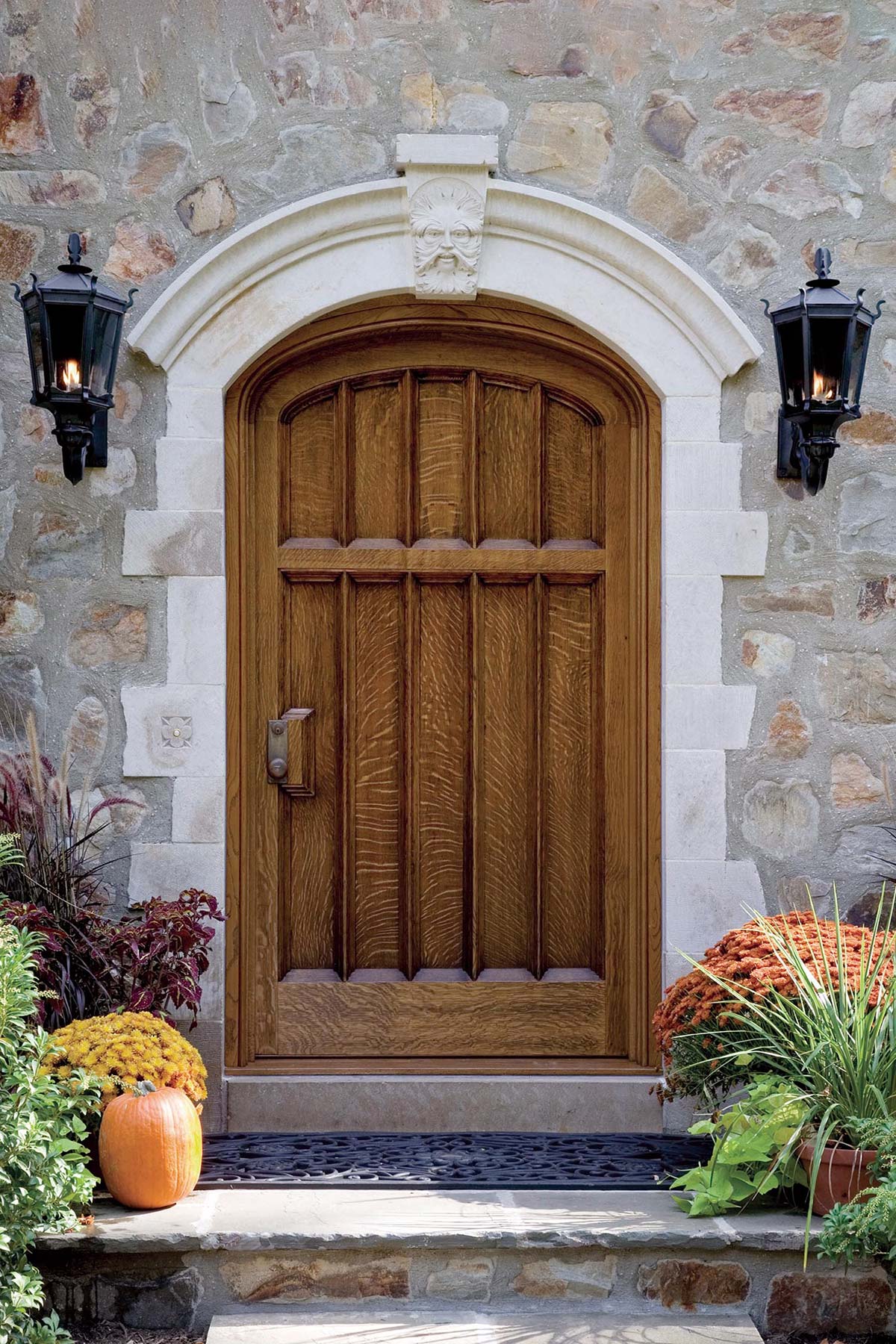 Elegant wooden door with stone wall, flanked by lanterns and autumn decor including pumpkins and mums.