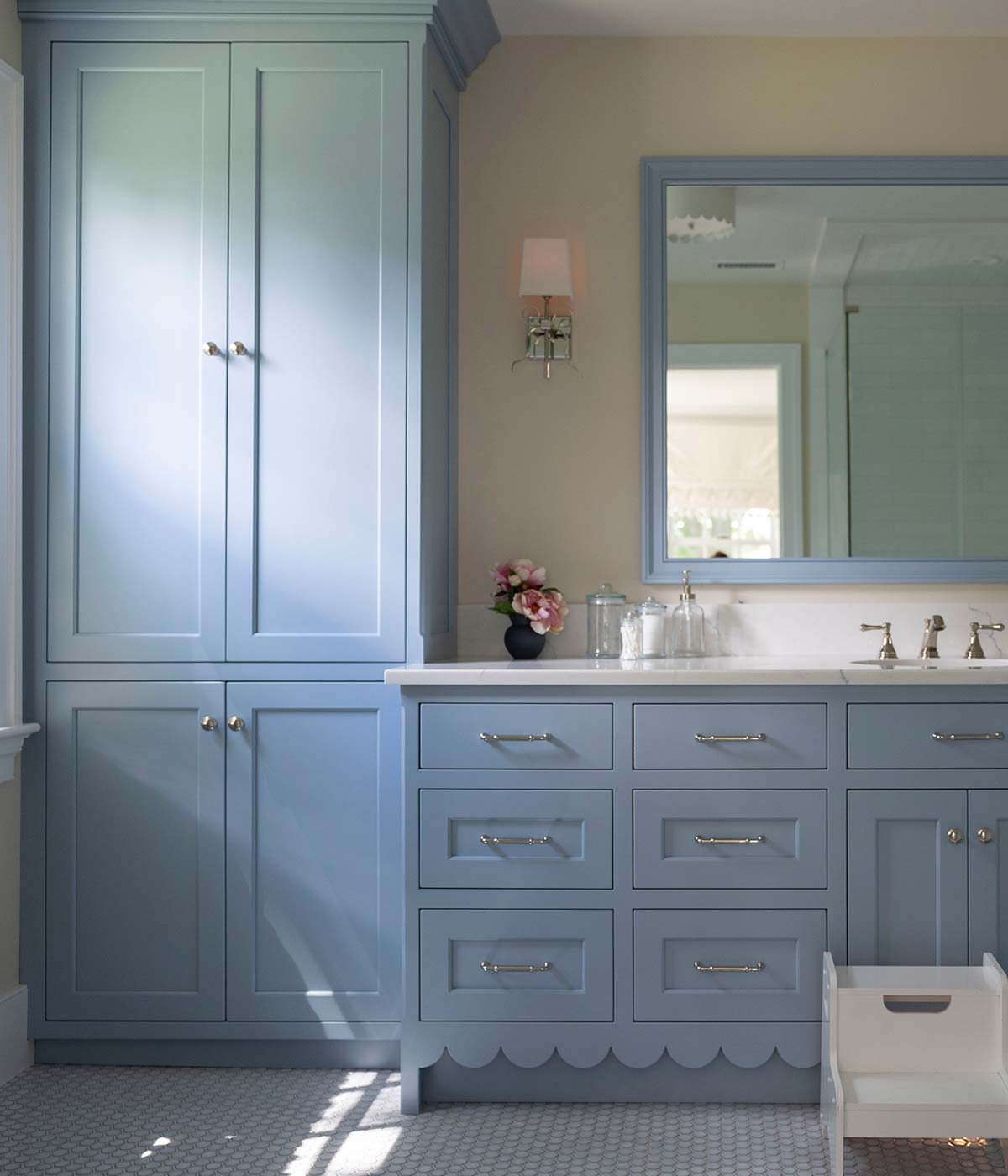 Blue bathroom cabinetry with scalloped edges, large mirror, and sink. Features a vase of flowers, white stool, and tiled floor with natural light.