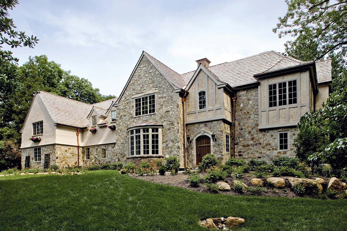 Grand stone house with steep gables, large windows, and lush landscaping, nestled among trees on a vibrant green lawn.
