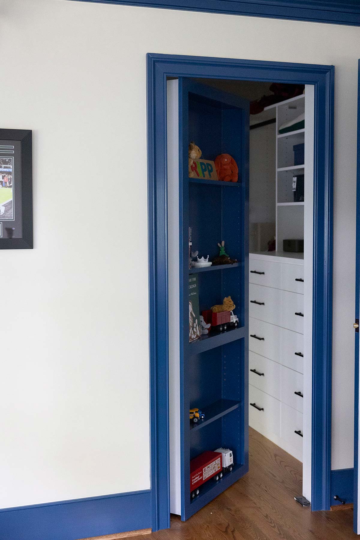 Blue bookshelf door revealing a closet with white drawers and toys. Includes a framed picture and wooden floor in a bright room.