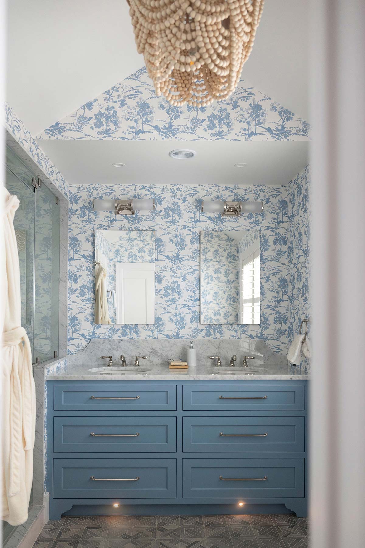 Blue bathroom with floral wallpaper, double sink, and beaded chandelier. Features mirrors, under-cabinet lighting, and tiled floor with natural light.