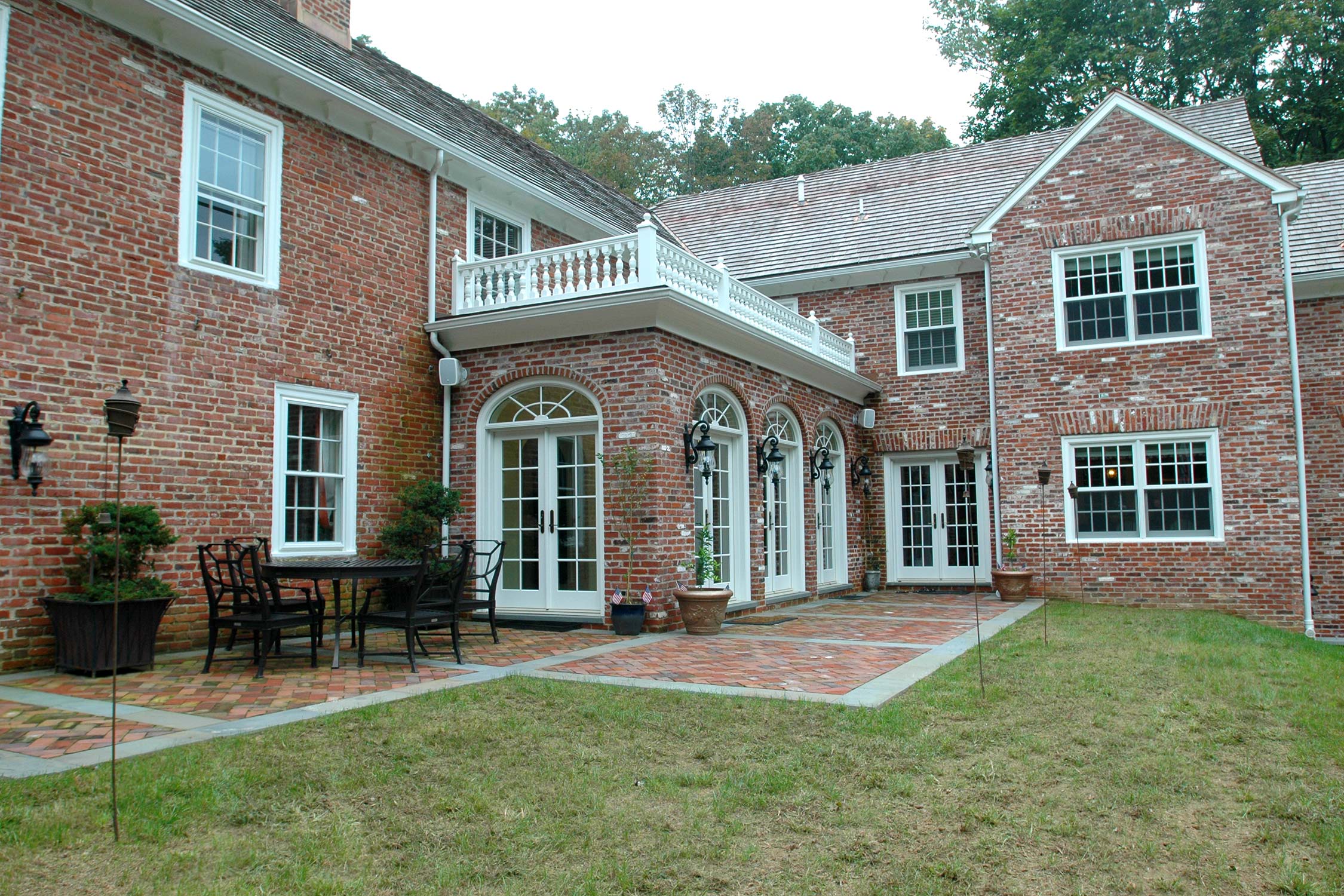 Elegant brick home with a balcony, arched windows, and French doors opening to a patio with outdoor seating and lush greenery.