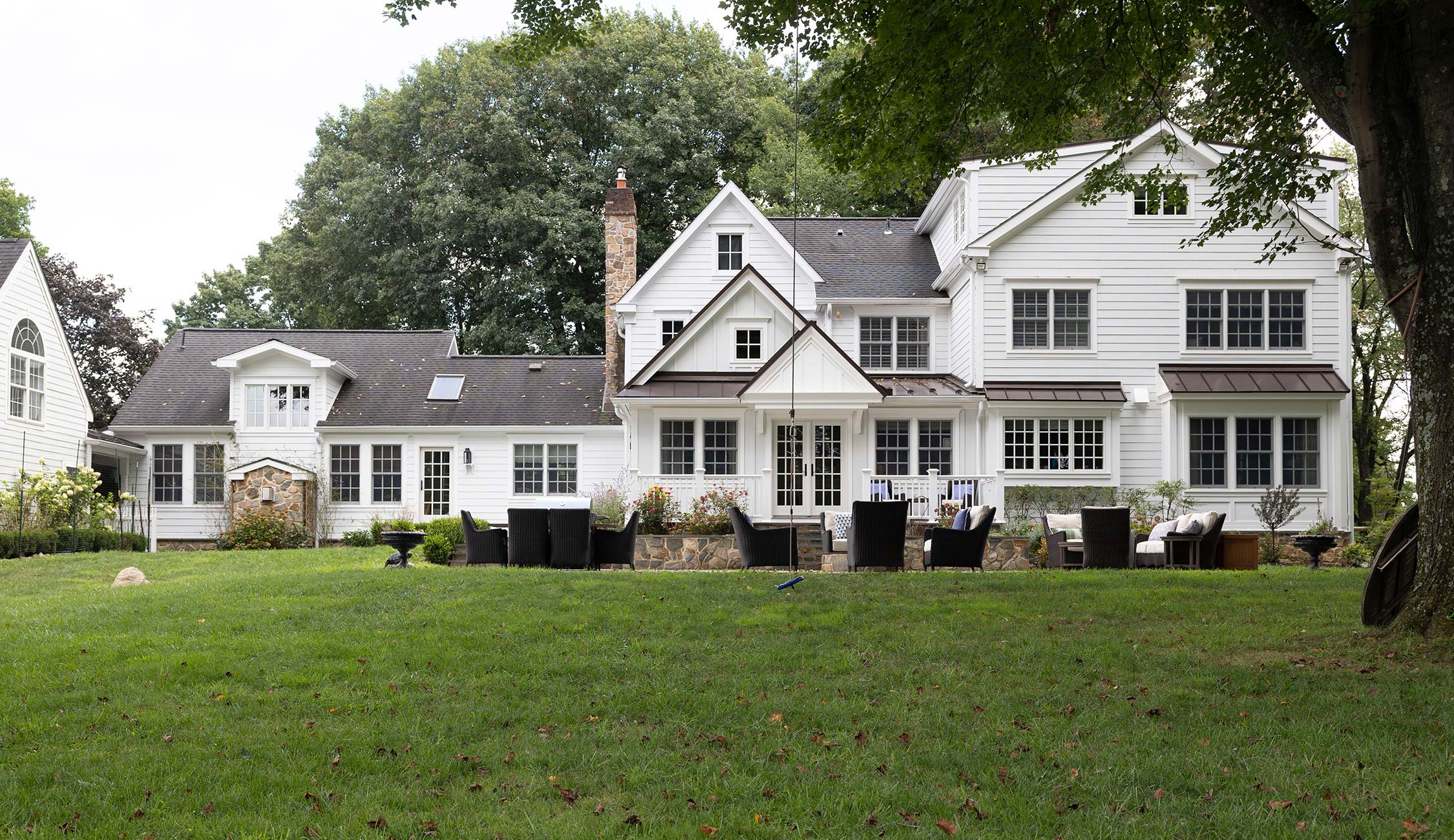 Spacious white house with stone accents, multiple windows, and a patio with wicker furniture and fire pit on a lush lawn.
