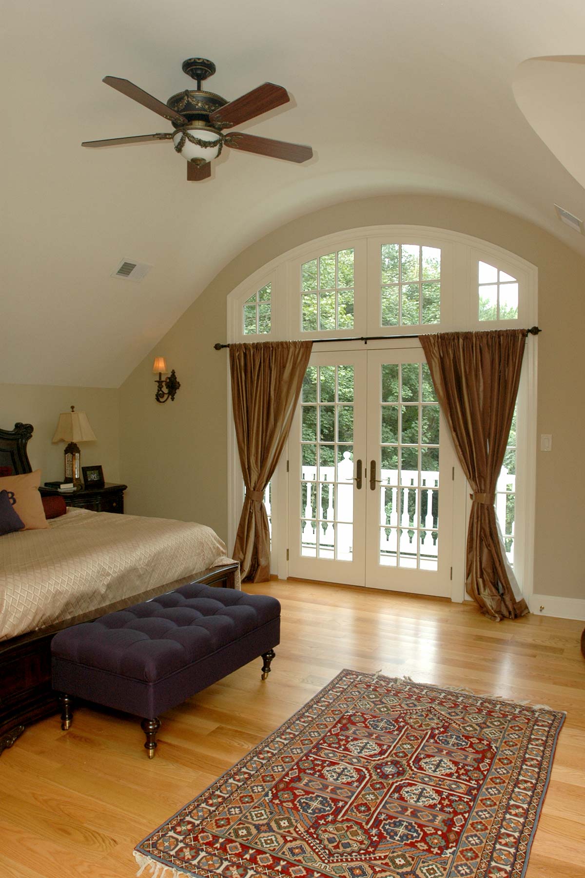 Serene bedroom with a ceiling fan, large arched windows, French doors to a balcony, and a cozy bed with a patterned rug on hardwood floors.