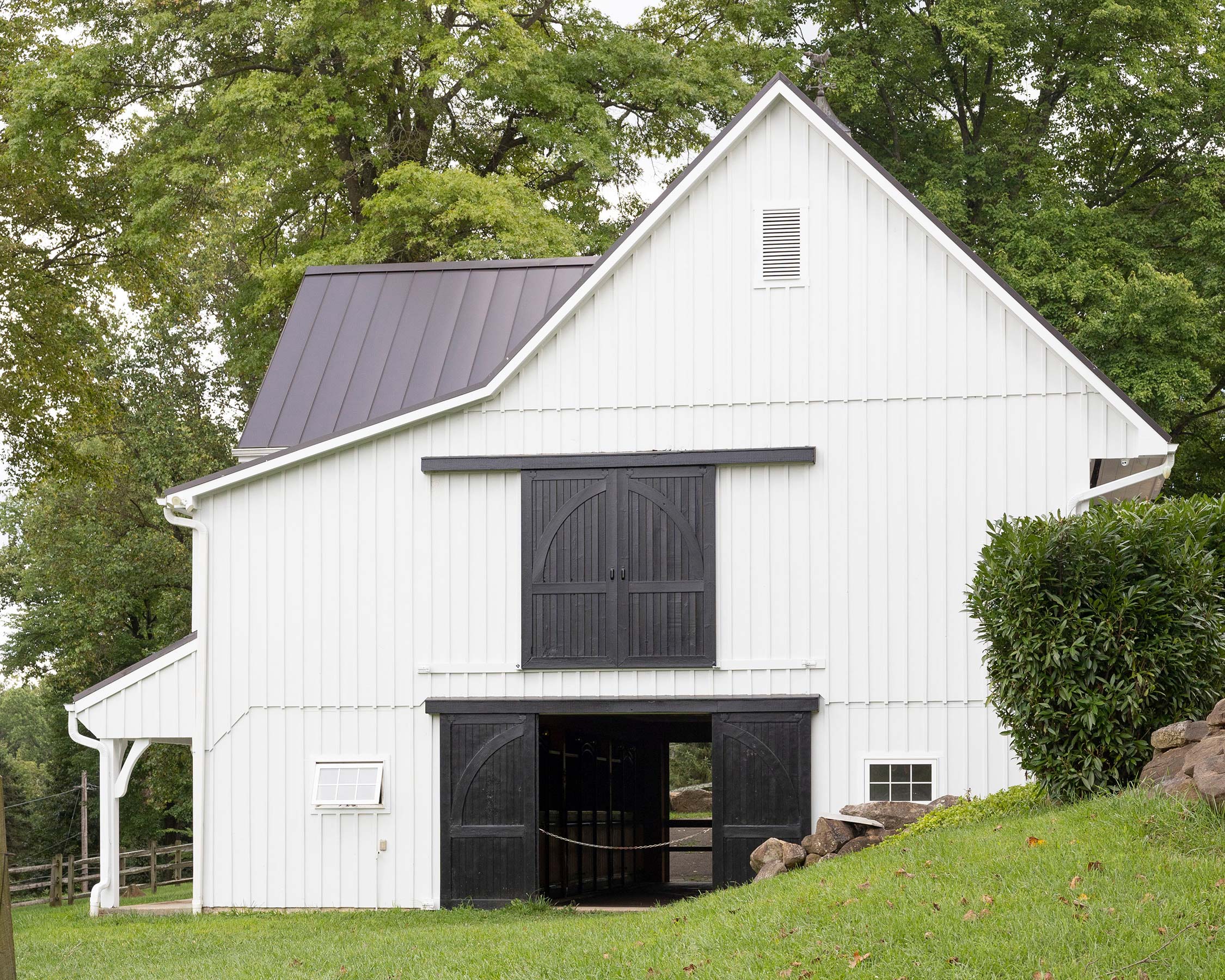 White barn with black double doors, metal roof, small windows, and surrounded by green lawn and trees on a hilly landscape.