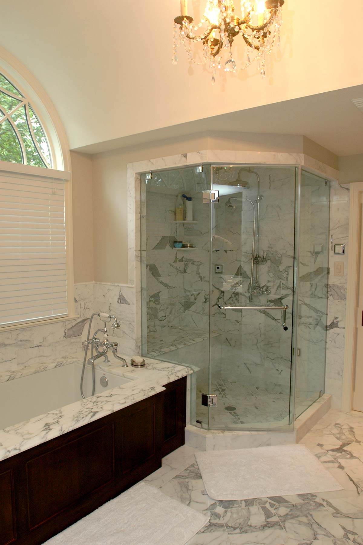 Luxurious bathroom with marble tub, glass shower, chandelier, and arched window. Features elegant dark cabinetry and a plush white rug.