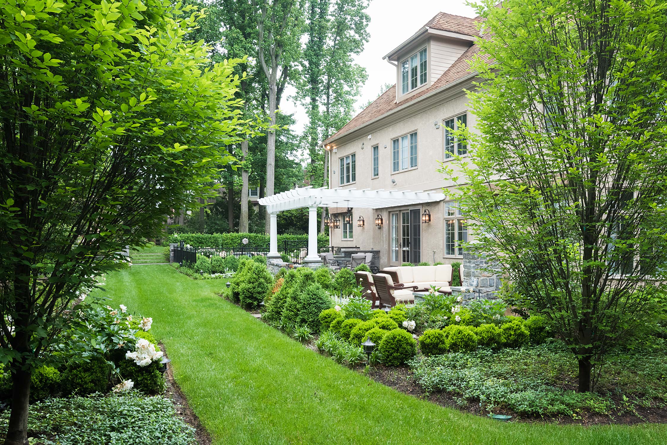 Elegant backyard with a beige house, white pergola, outdoor seating, and lush gardens framed by tall trees.