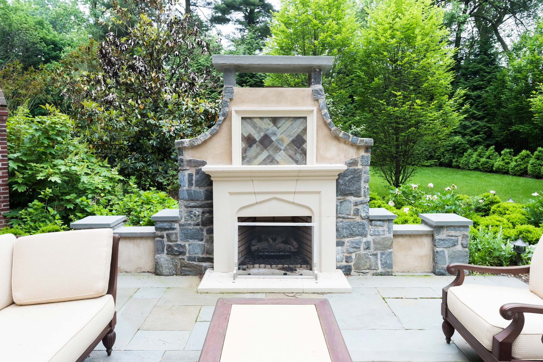 Cozy outdoor fireplace with stone and stucco design, surrounded by lush greenery and comfortable seating on a stone patio.