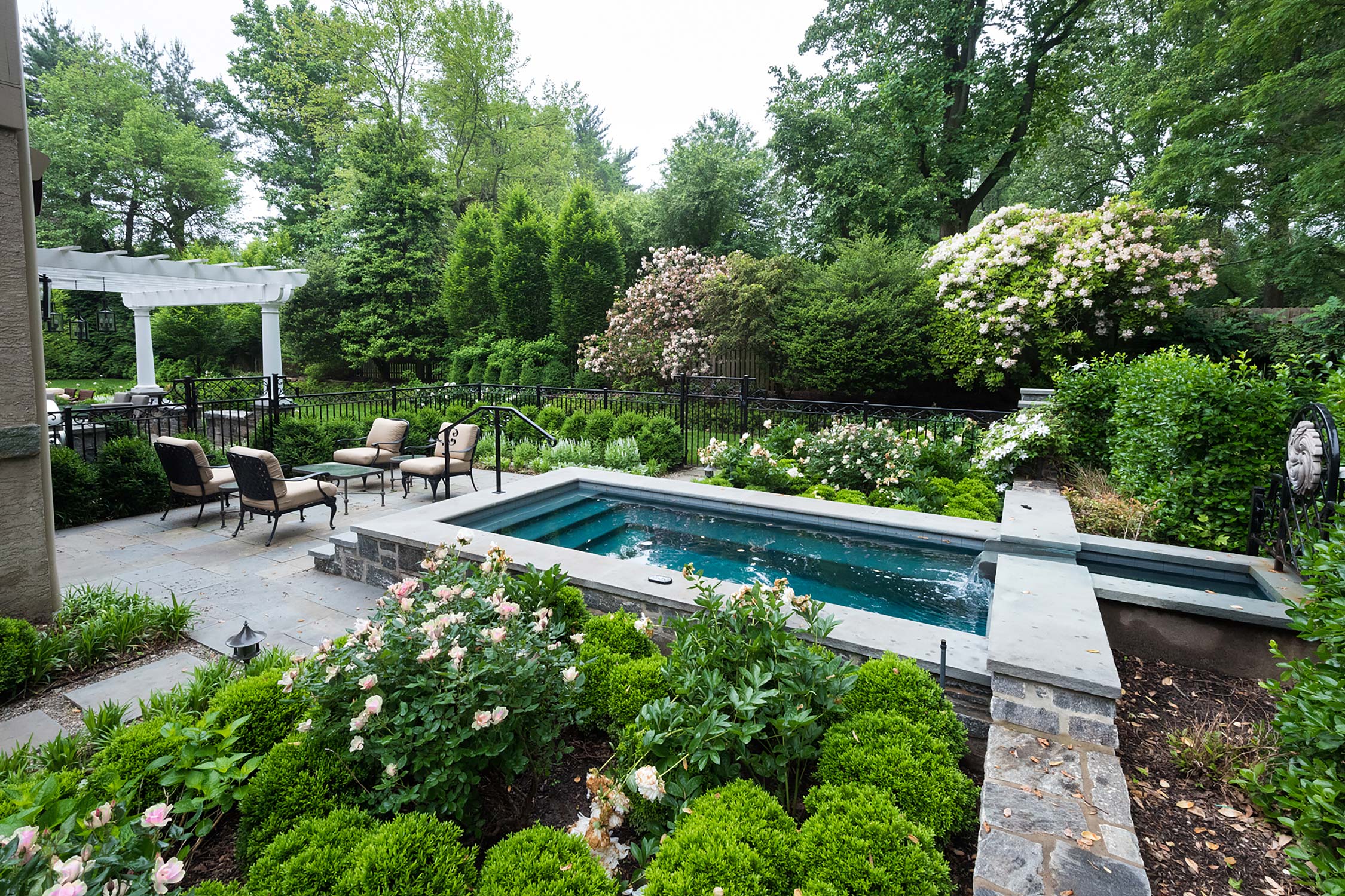 Tranquil backyard pool with stone patio, white pergola, and vibrant gardens surrounded by lush trees and seating area.
