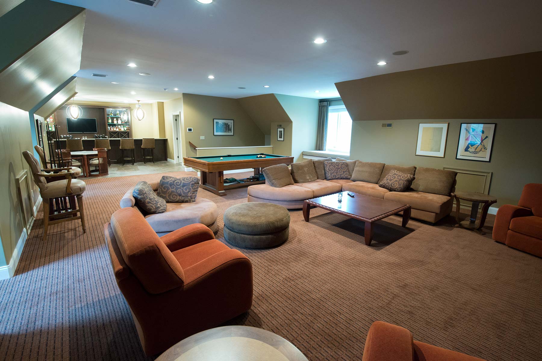 Cozy game room with pool table, bar area, plush seating, and modern lighting on warm carpeted floors.