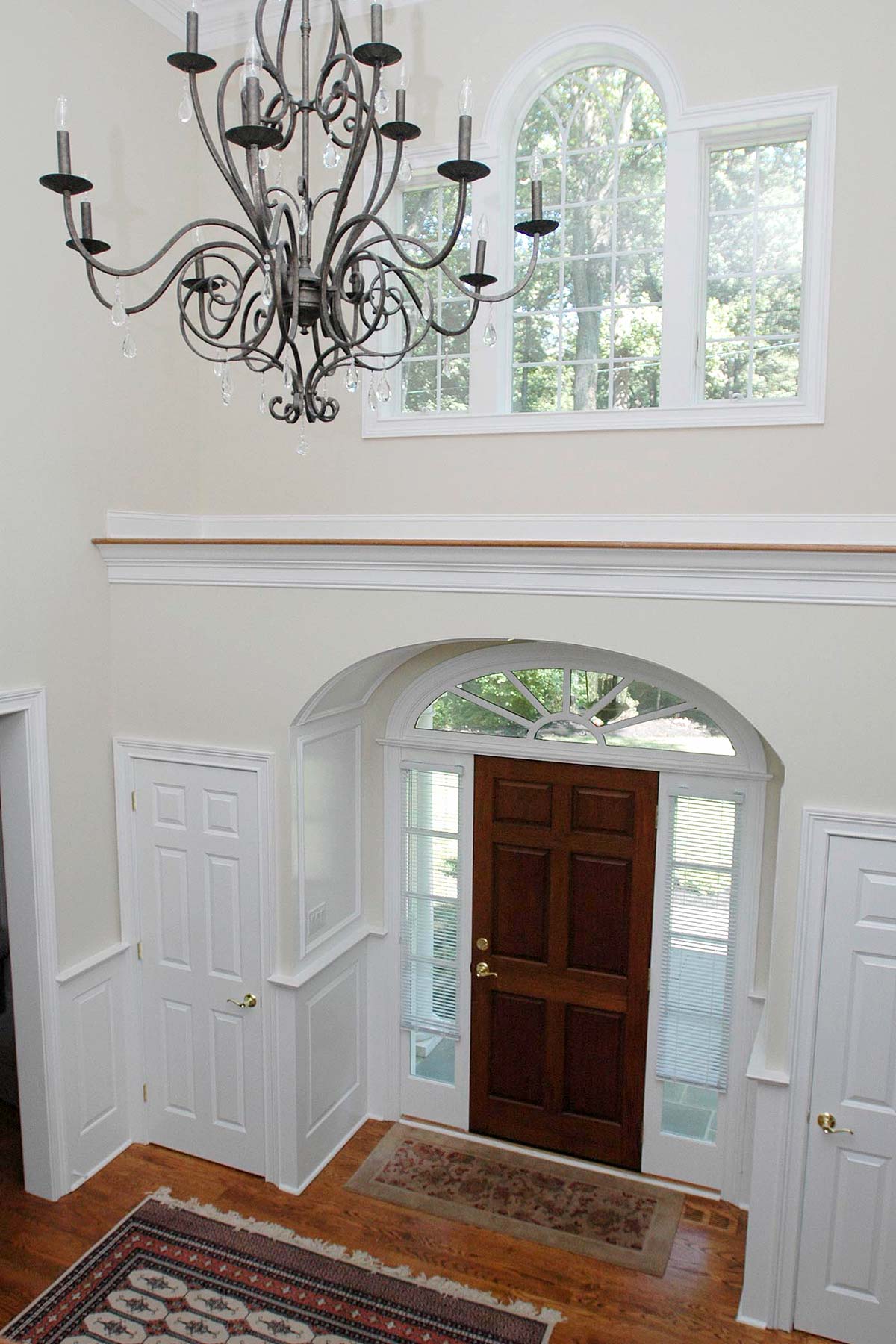 Grand foyer with ornate chandelier, arched windows, wooden door, and a patterned rug on polished wood floors.