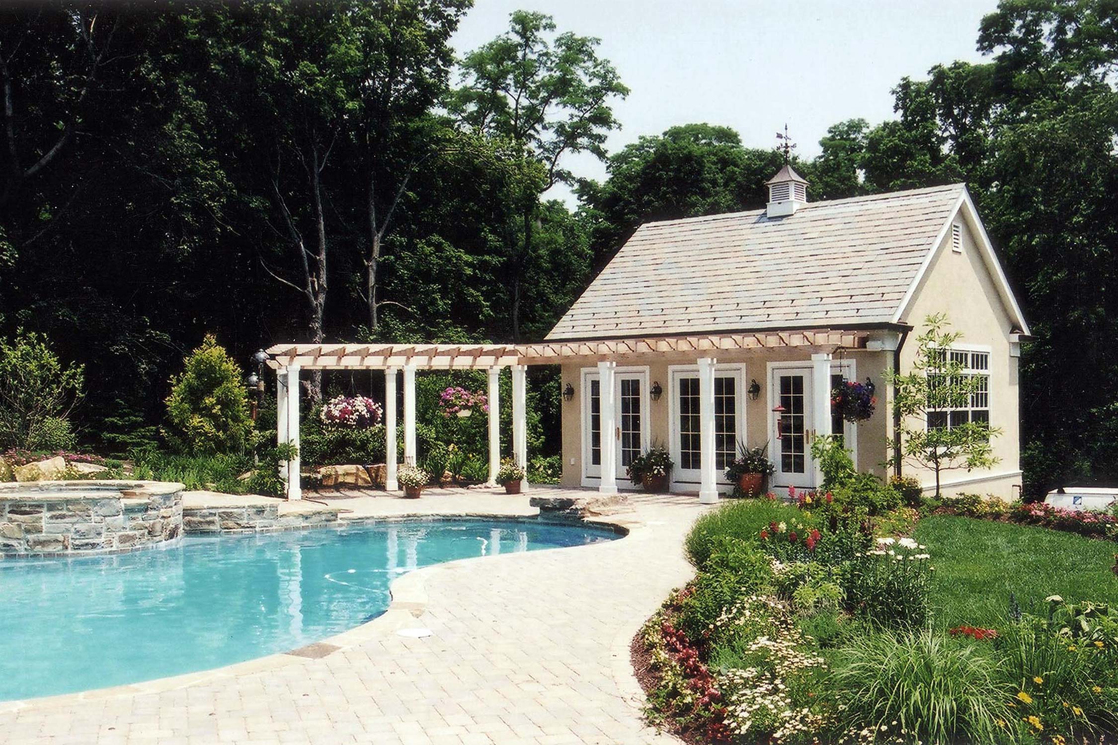 Charming poolside cottage with white pergola, surrounded by vibrant gardens and lush trees on a sunny day.