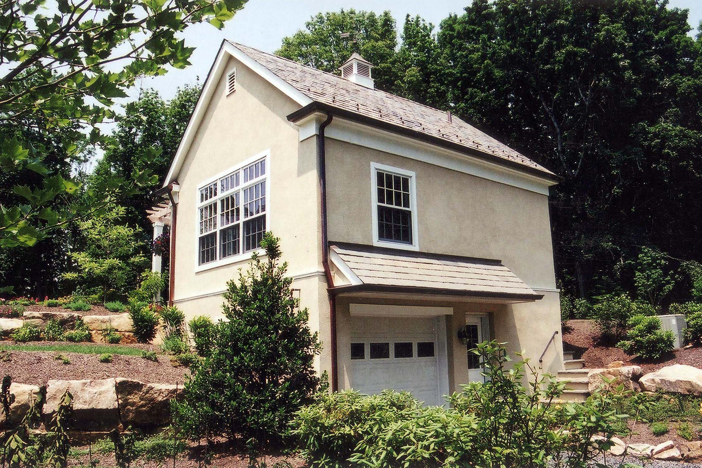 Charming small house with beige exterior, large windows, and a garage, nestled among lush greenery and rocks on a sunny day.