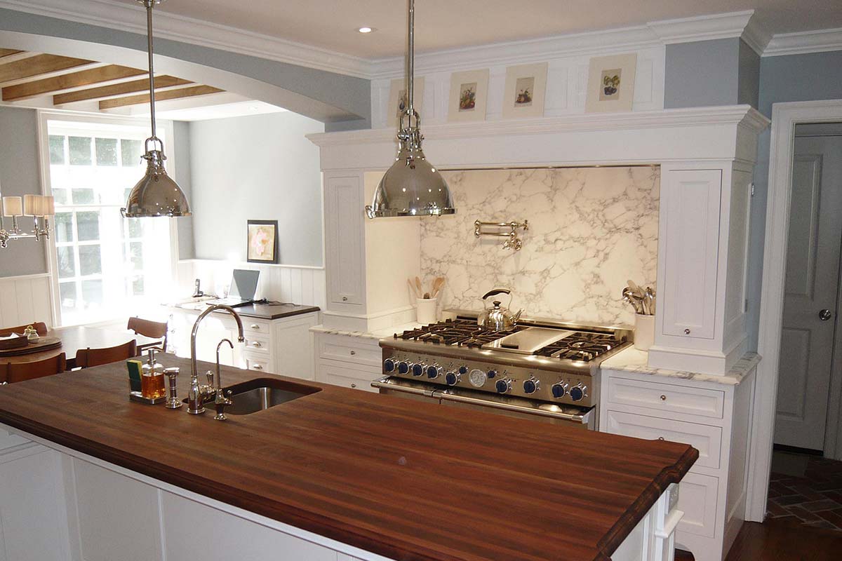Modern kitchen with wooden countertop, stainless steel stove, and elegant pendant lights, featuring white cabinets and marble backsplash.