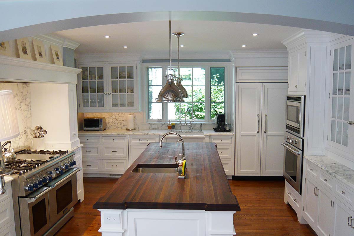 Bright kitchen with wooden island, stainless steel stove, and white cabinets, featuring large windows and elegant pendant lights.