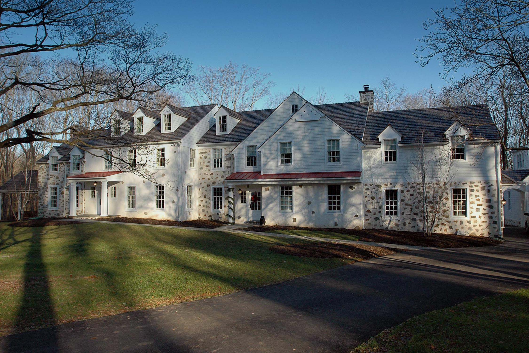 Elegant white house with stone accents, dormer windows, and red roof, surrounded by trees and a well-maintained lawn on a sunny day.