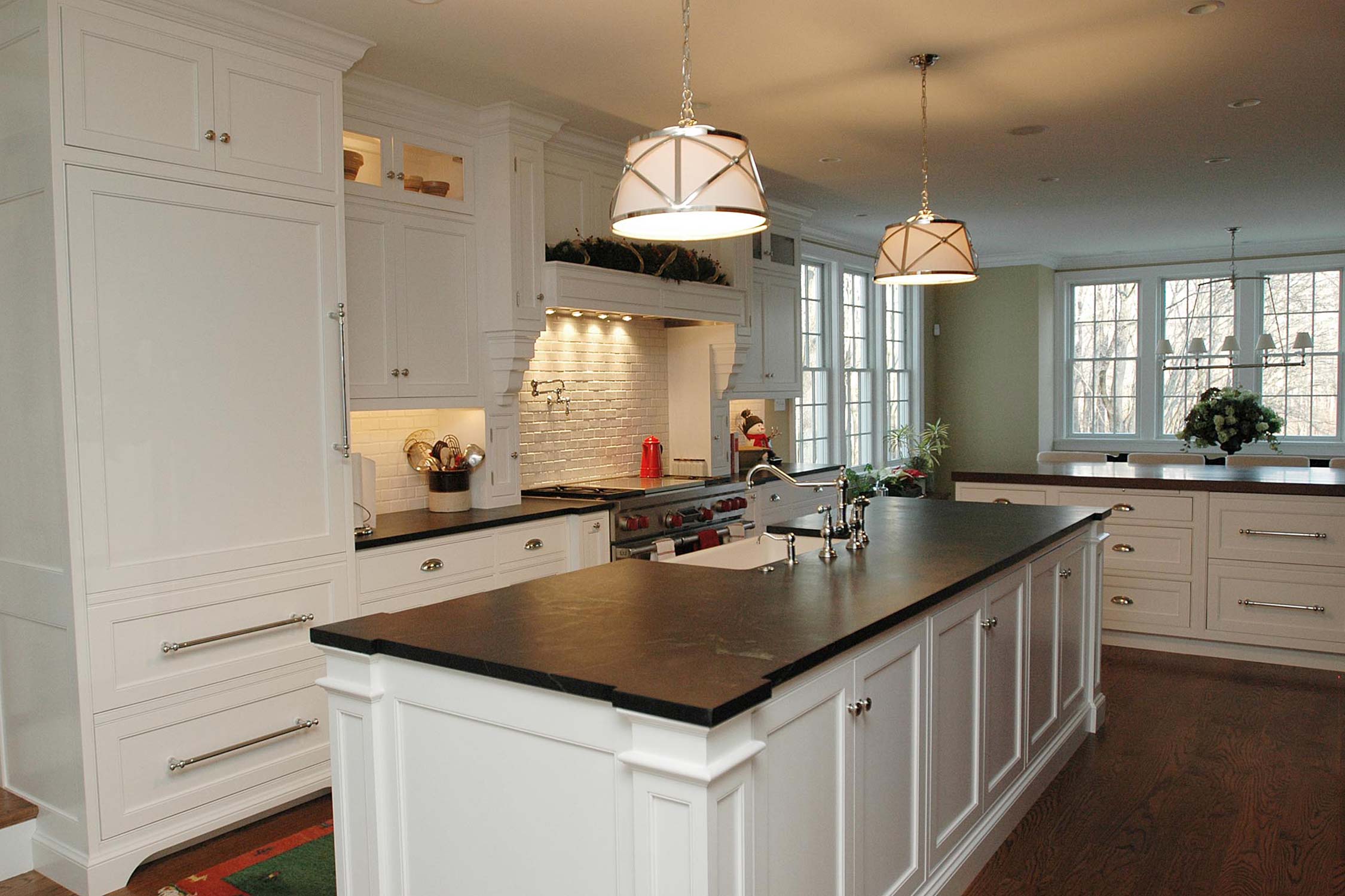 Modern kitchen with dark granite island, white cabinets, and pendant lights, featuring a brick backsplash and large windows.