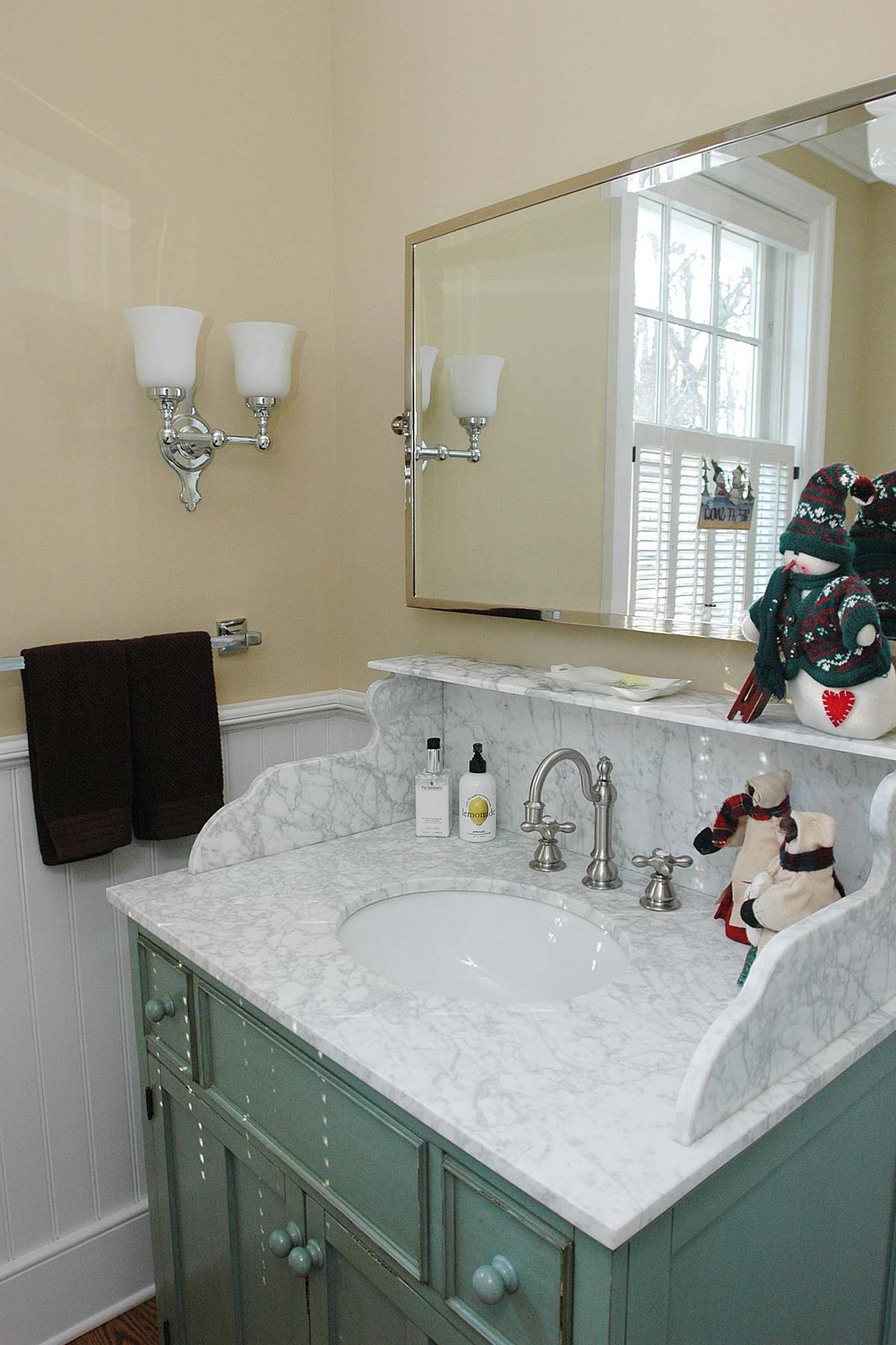 Elegant bathroom with marble sink, teal vanity, and wall sconces, decorated with holiday snowmen near a bright window.