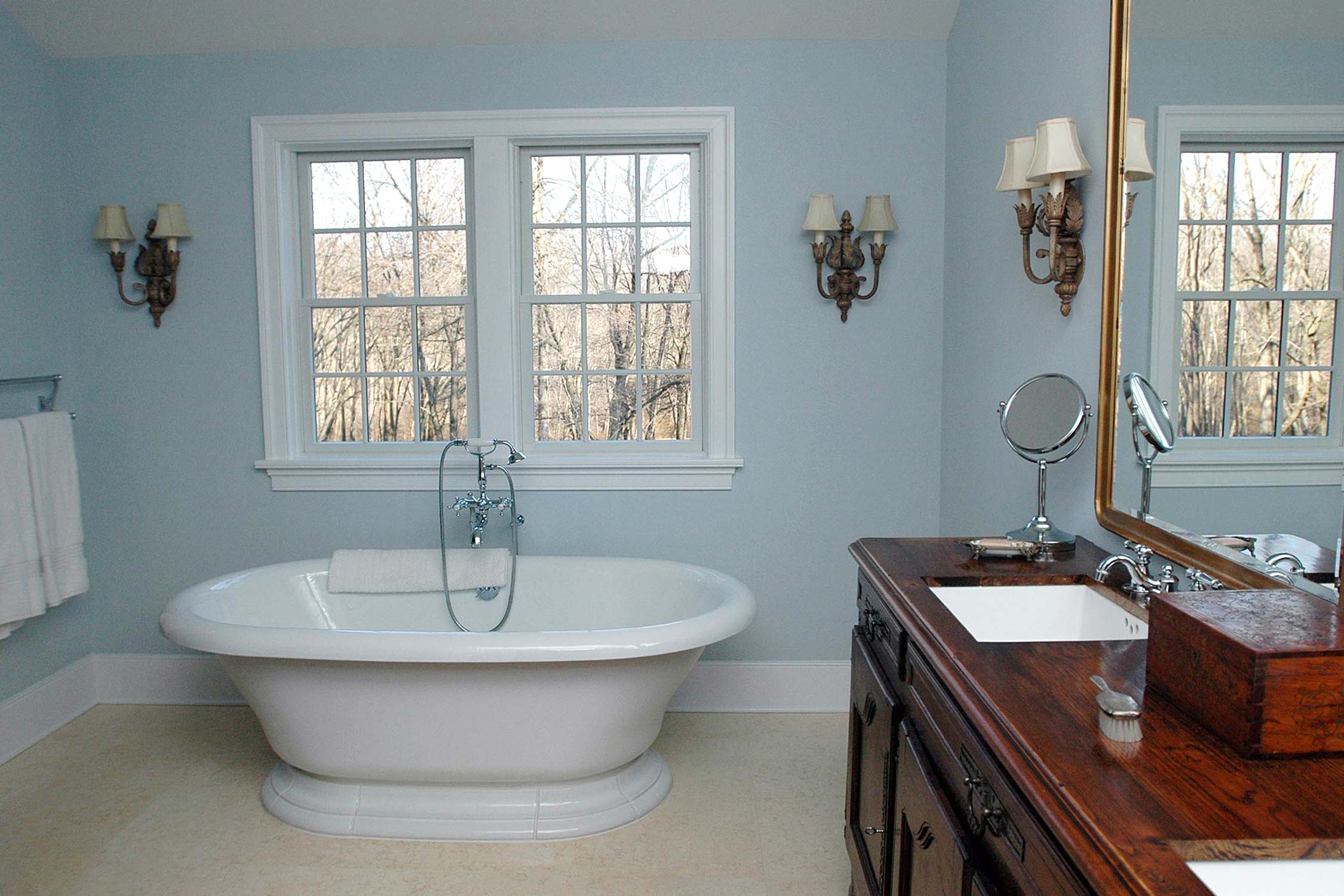 Serene bathroom with a freestanding bathtub, wooden vanity, and large windows, featuring elegant wall sconces and soft lighting.