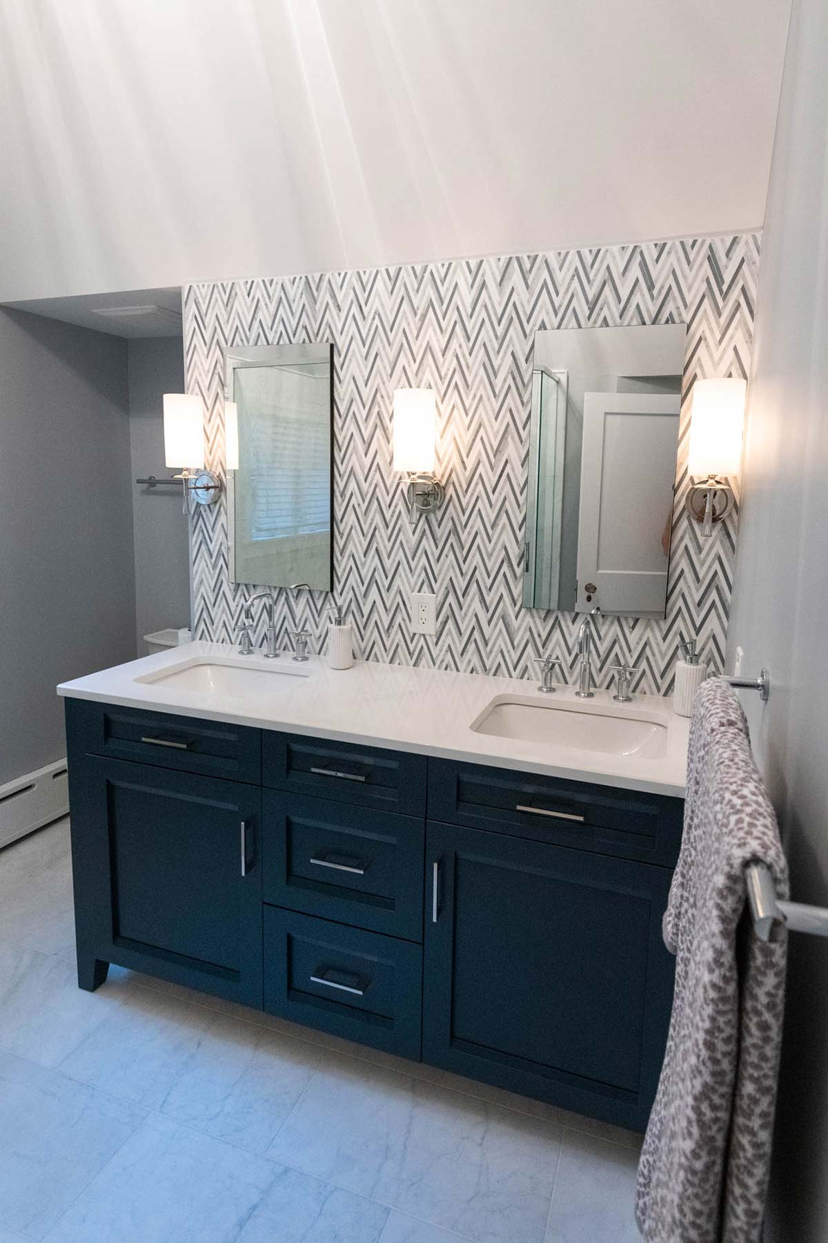 Modern bathroom with dual sinks, dark blue vanity, and chevron tile backsplash, featuring elegant lighting and marble flooring.