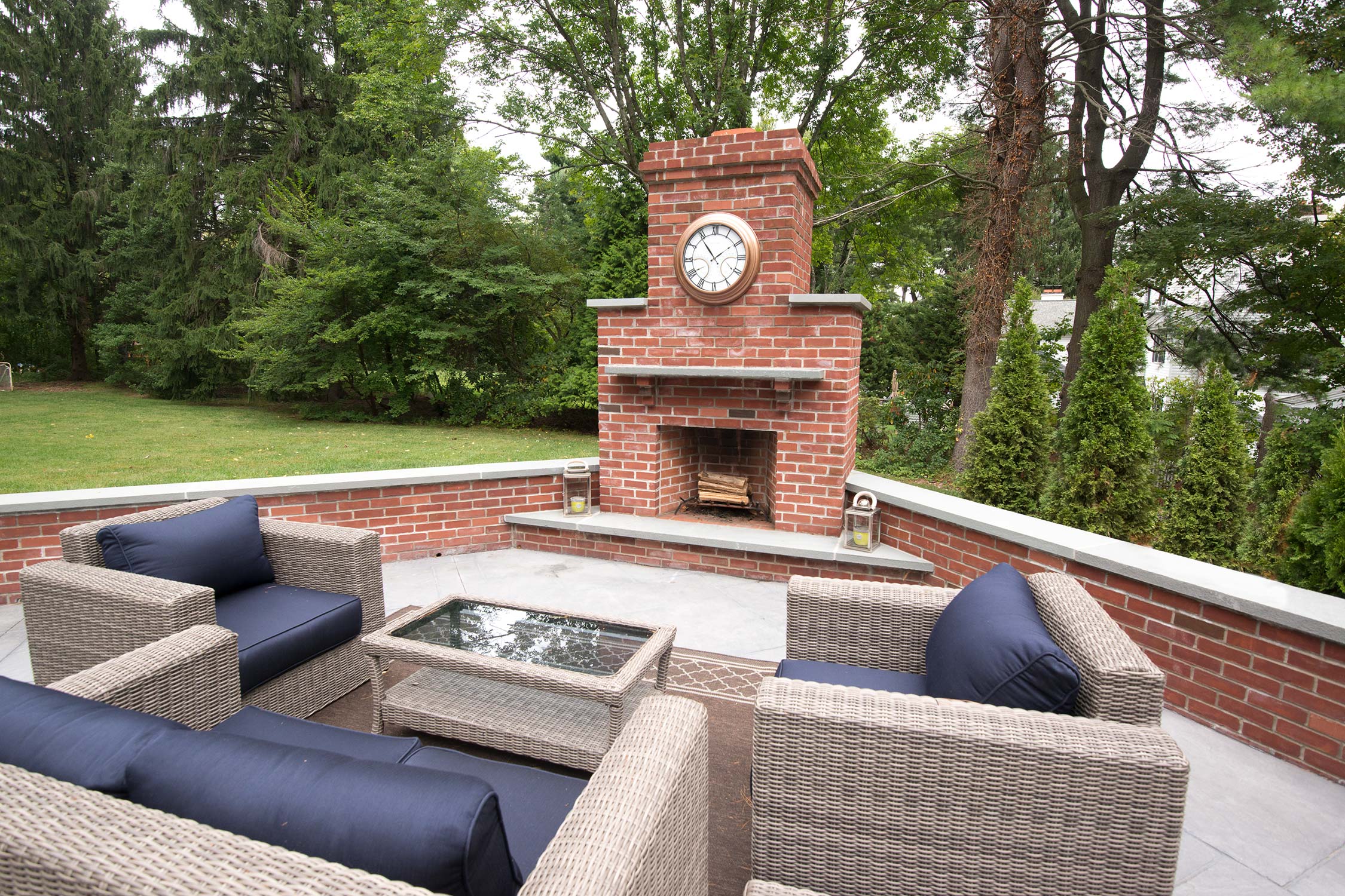Cozy outdoor patio with a brick fireplace, clock, and wicker furniture, surrounded by lush greenery on a serene day.