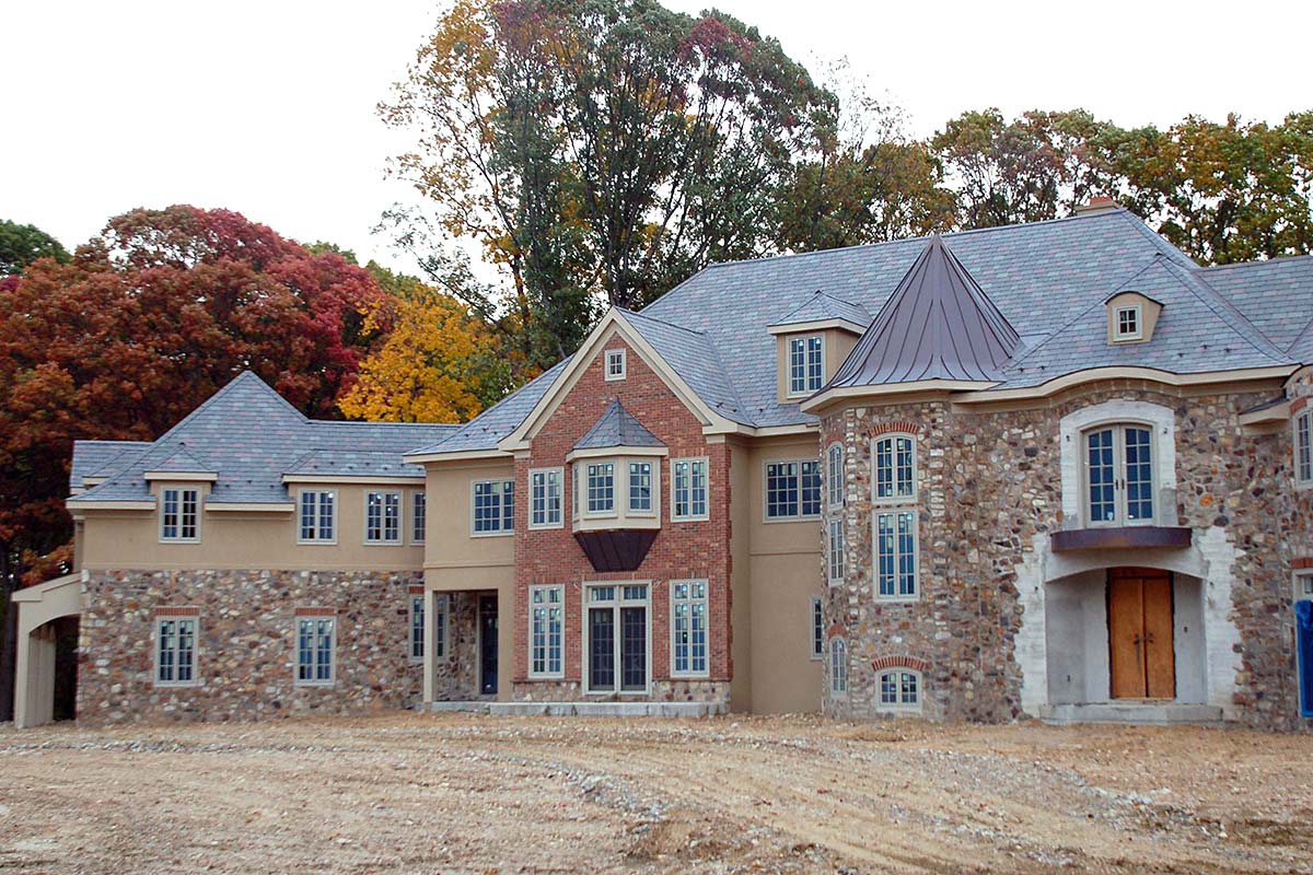Grand house with stone and brick exterior, steep roofs, and large windows, set against a backdrop of colorful autumn trees.