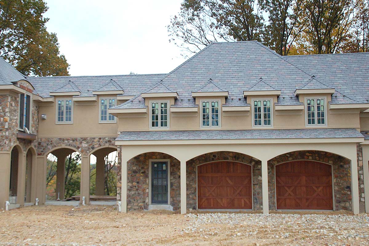 Elegant house with stone and stucco exterior, arched garage doors, and dormer windows, set against autumn trees on a clear day.