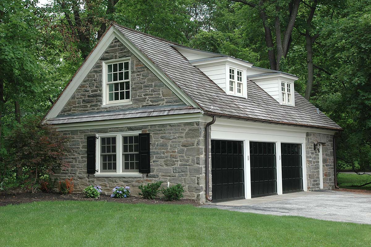Charming stone garage with double doors and dormer windows, surrounded by lush greenery and vibrant flowers.