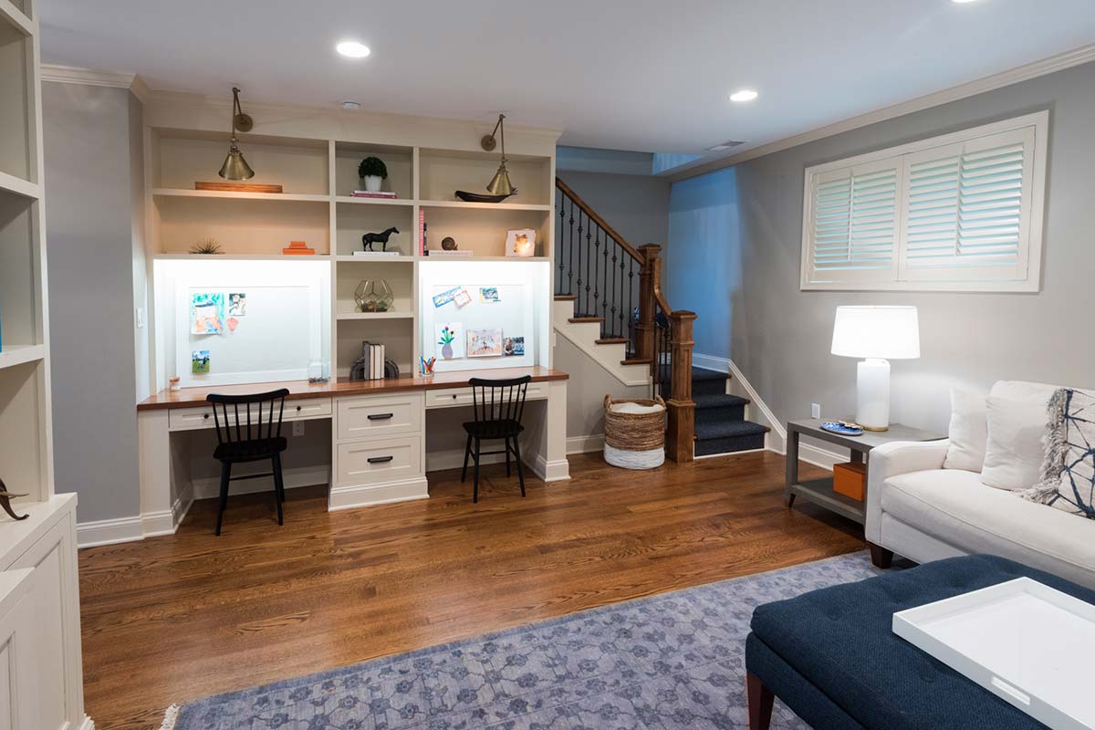 Cozy room with dual desks, wooden flooring, and a staircase, featuring a sofa, ottoman, and stylish decor near a window.