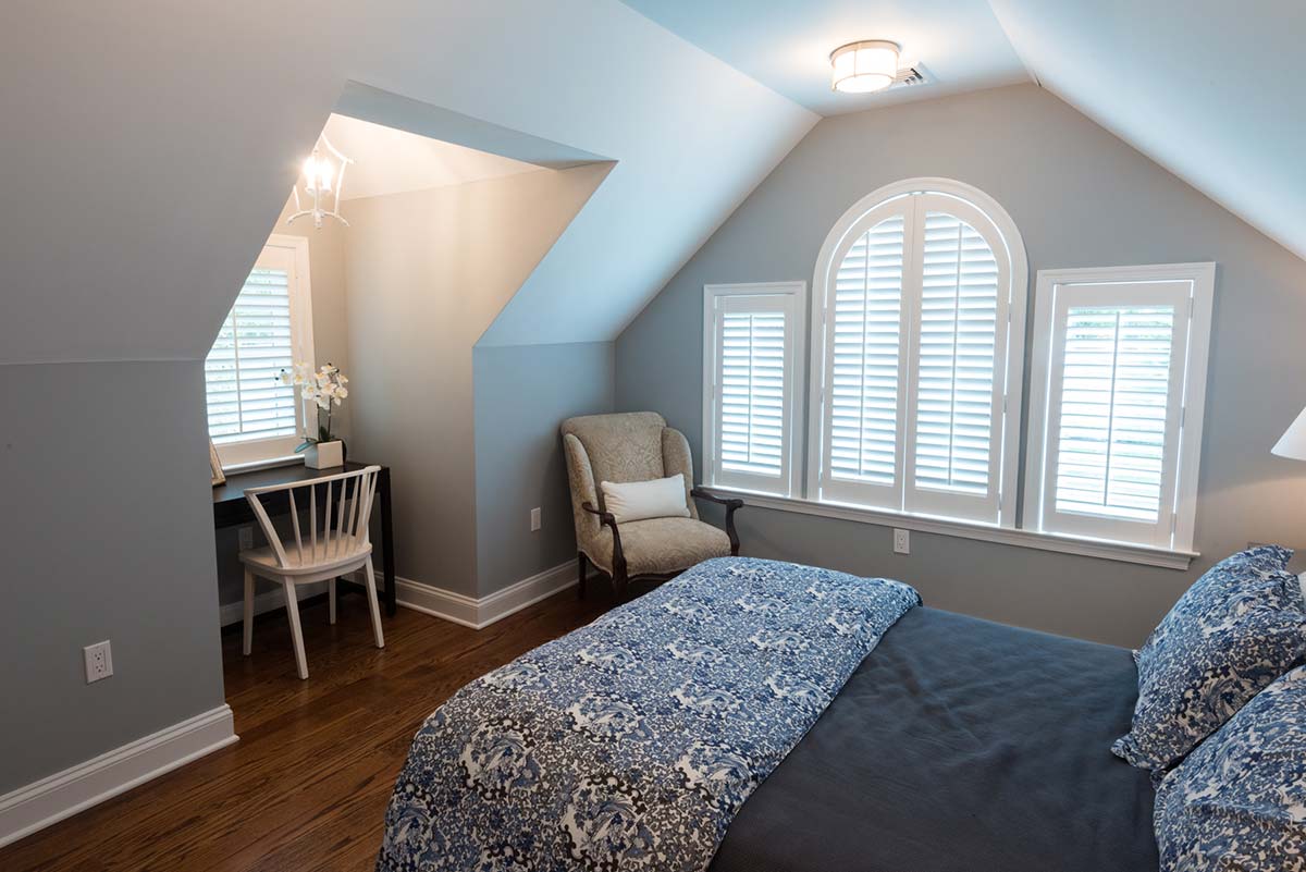 Cozy bedroom with slanted ceiling, wooden floor, and blue patterned bedding, featuring a chair, desk, and arched windows.