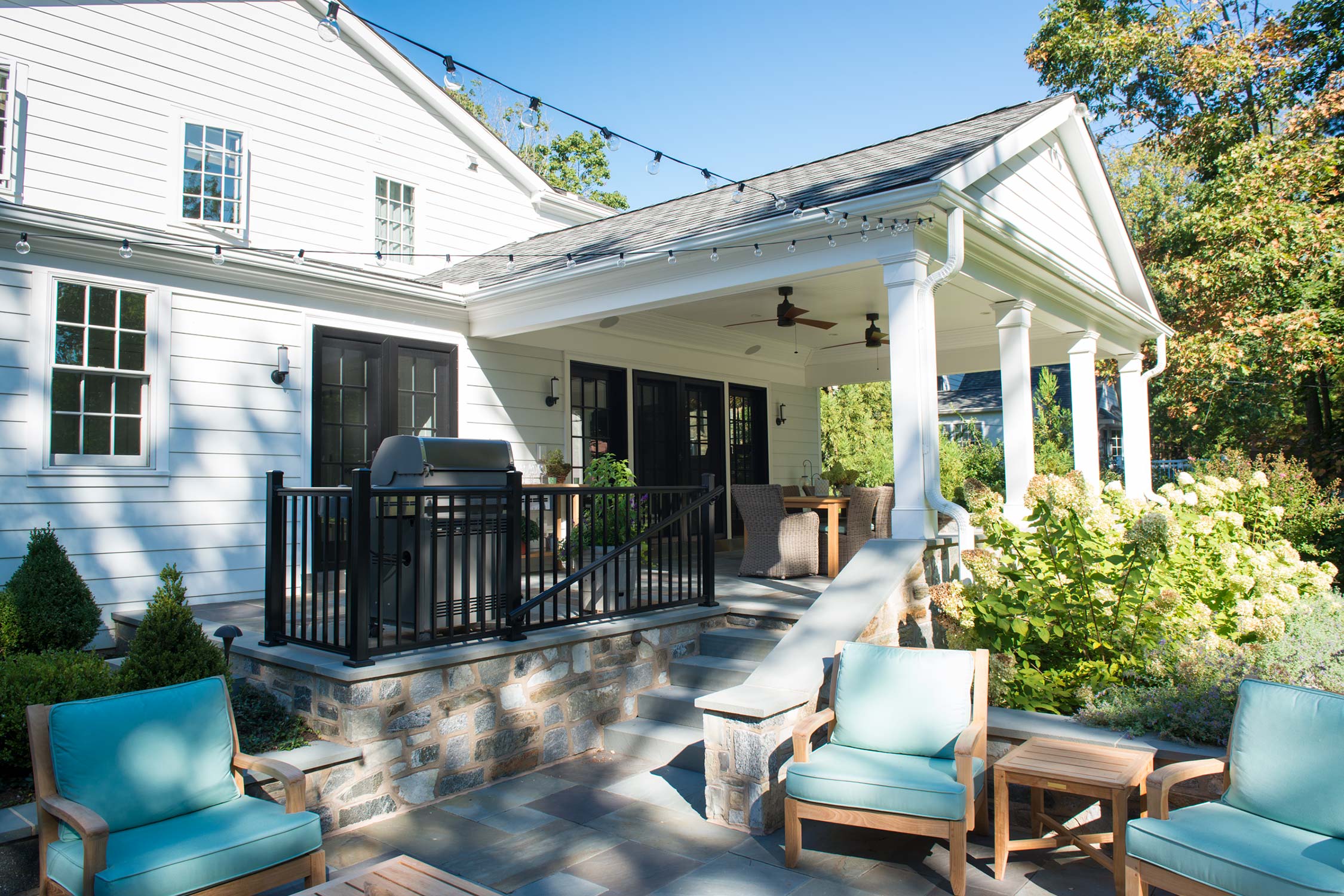 Cozy outdoor patio with white house, string lights, blue chairs, and lush greenery under a clear sky.