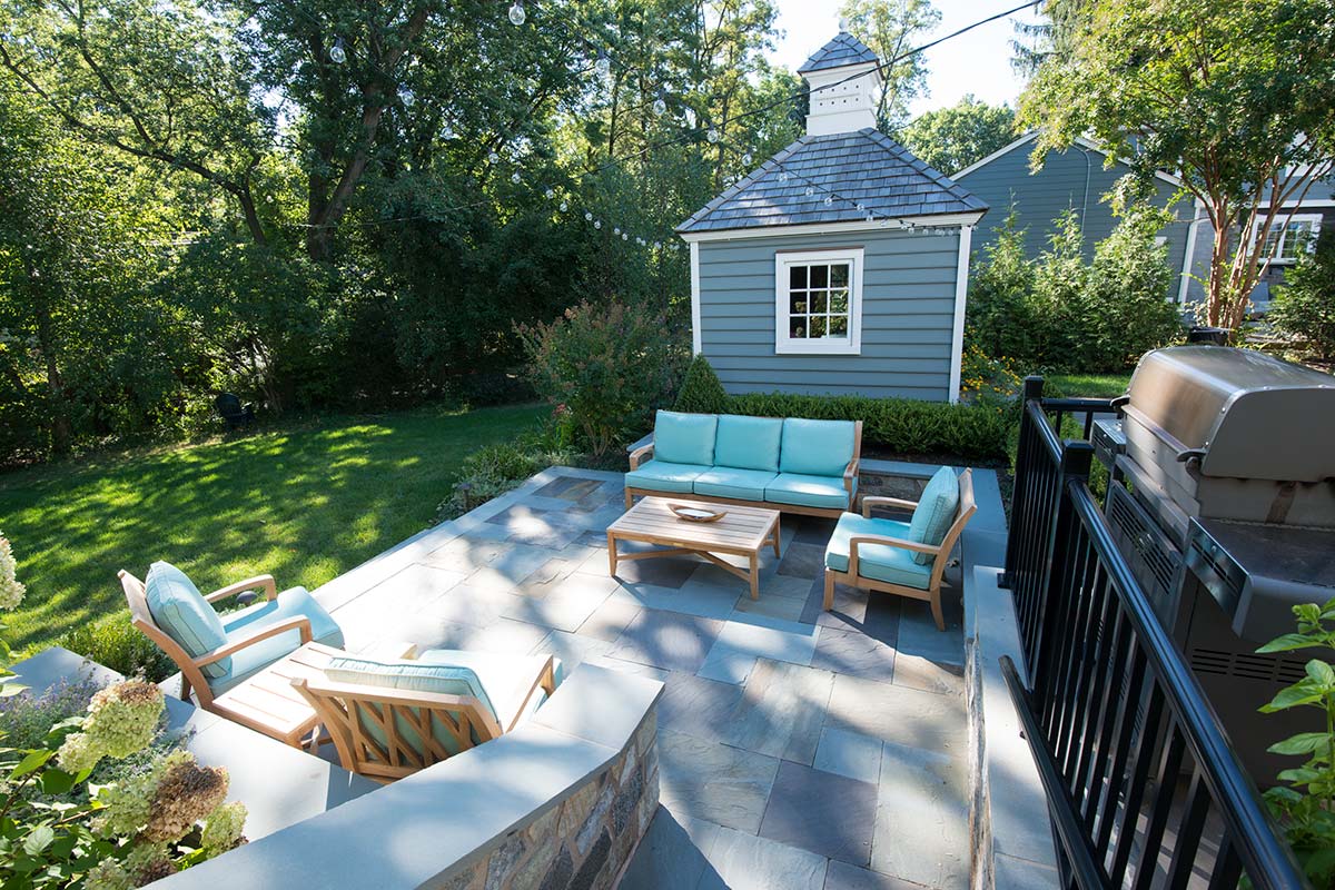 Serene backyard patio with turquoise seating, wooden tables, outdoor grill, blue shed, and lush green surroundings under sunny skies.