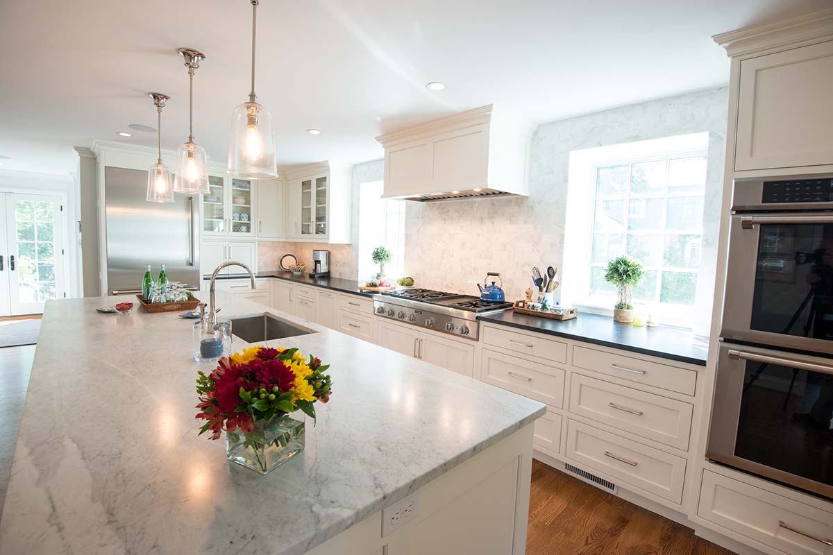 Bright modern kitchen with marble island, colorful flowers, white cabinets, stainless steel appliances, and natural light from large windows.