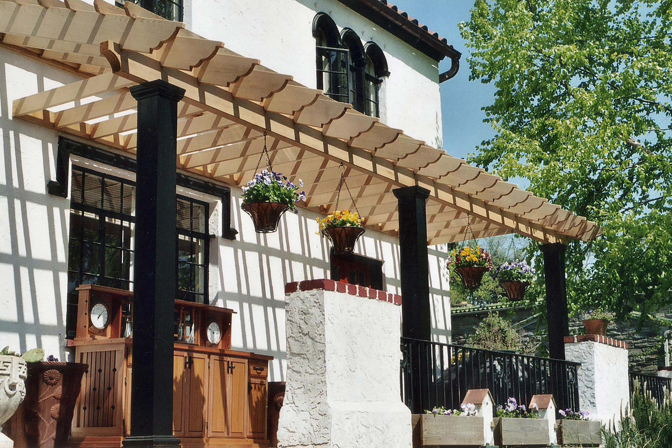 Charming patio with wooden pergola, hanging flower baskets, black columns, and white stucco house under a clear blue sky.