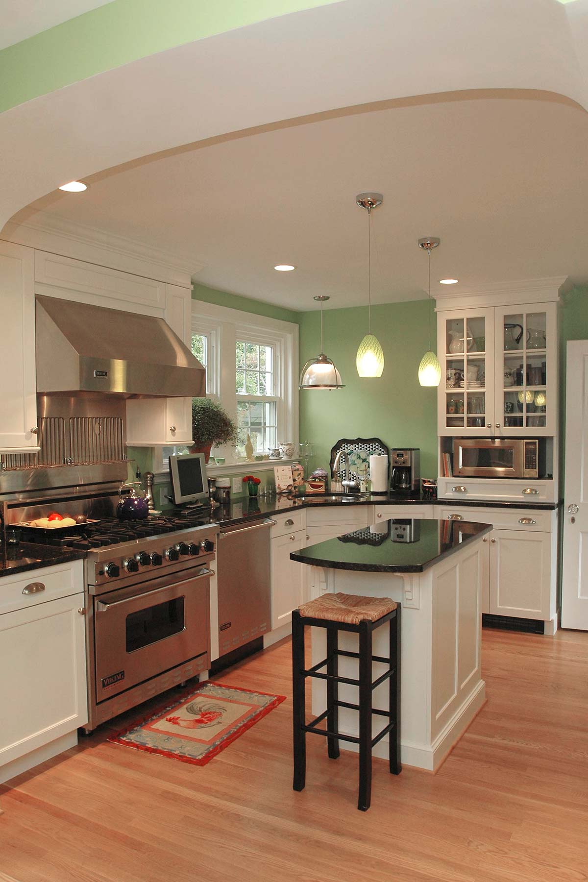 Modern kitchen with green accents, curved ceiling, white cabinets, black granite island, and hardwood floors under soft lighting.