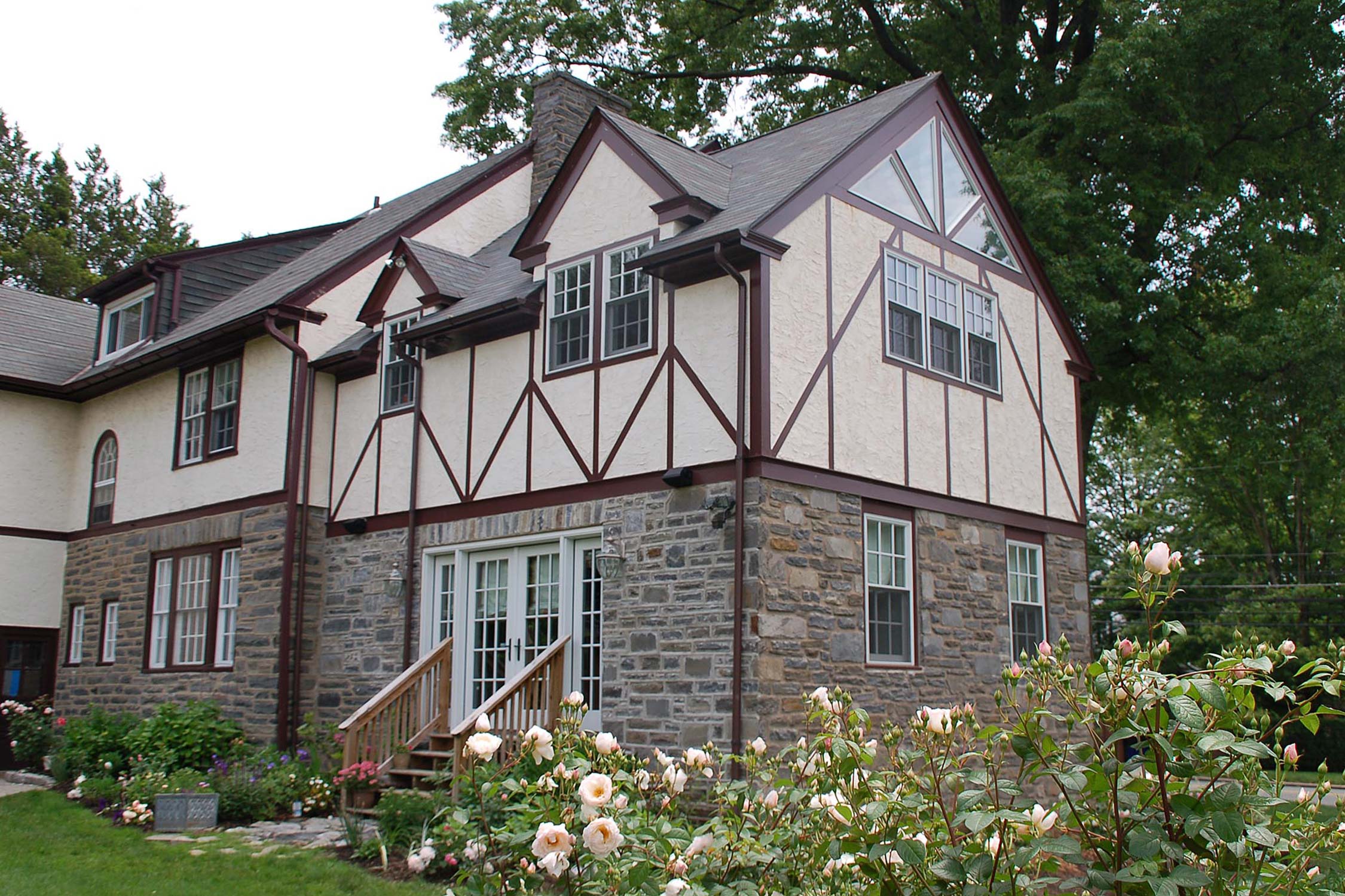 Elegant Tudor-style home with stone base, timber framing, and lush rose garden under a cloudy sky.