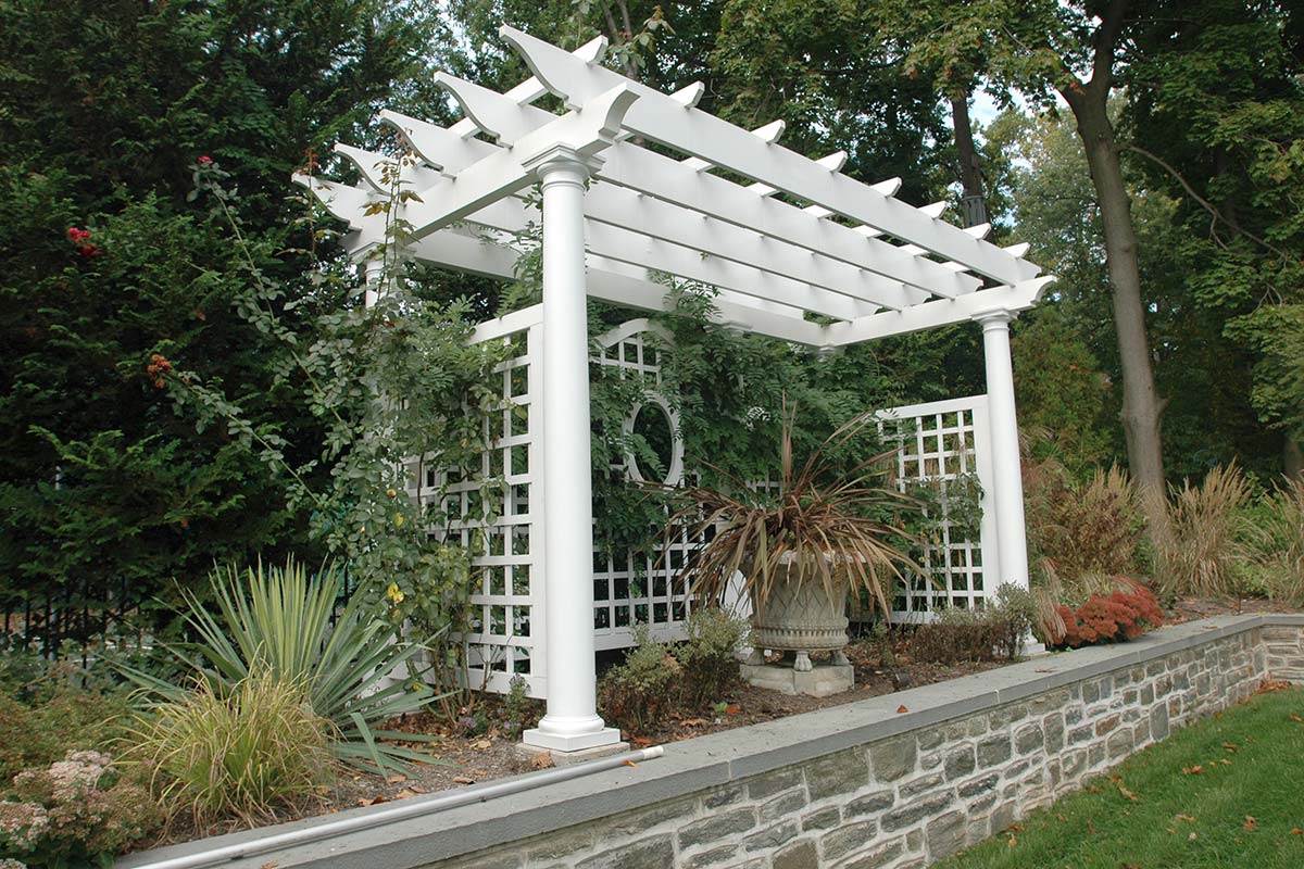 Elegant garden pergola with white columns, lush greenery, and stone wall, surrounded by vibrant plants and trees.