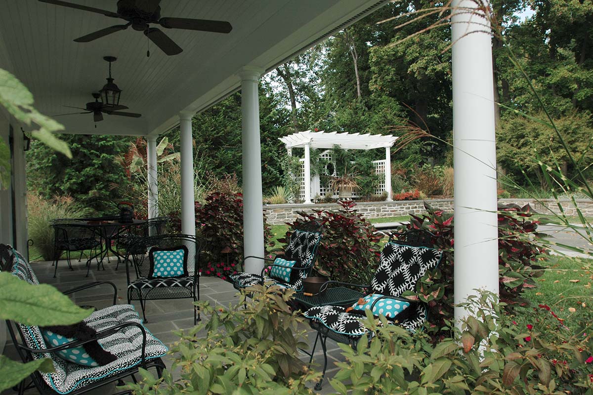 Cozy porch with white columns, ceiling fans, patterned chairs, and lush greenery, overlooking a garden pergola.