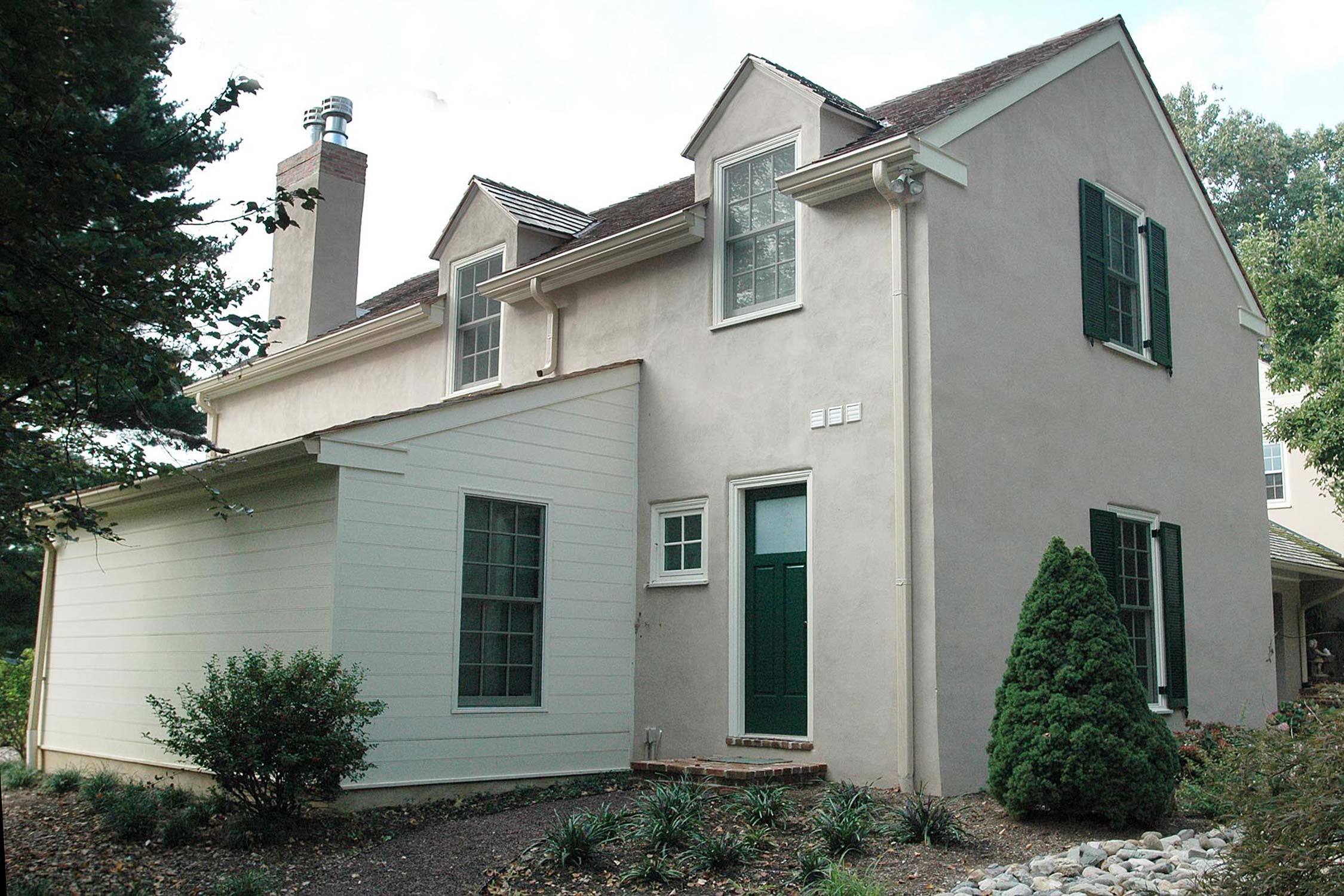 Charming home with stucco exterior, green door, stone chimney, and lush landscaping under a cloudy sky.