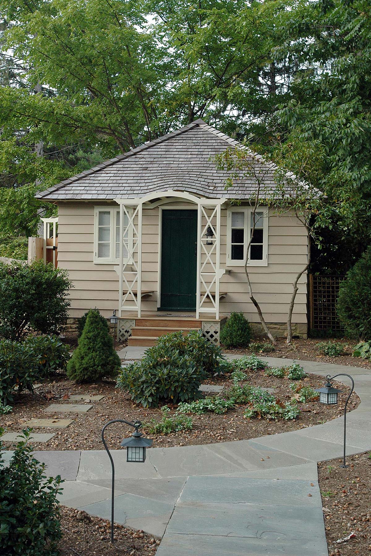 Cozy cottage with green door and white trim, nestled among lush trees and a stone pathway with garden lights.